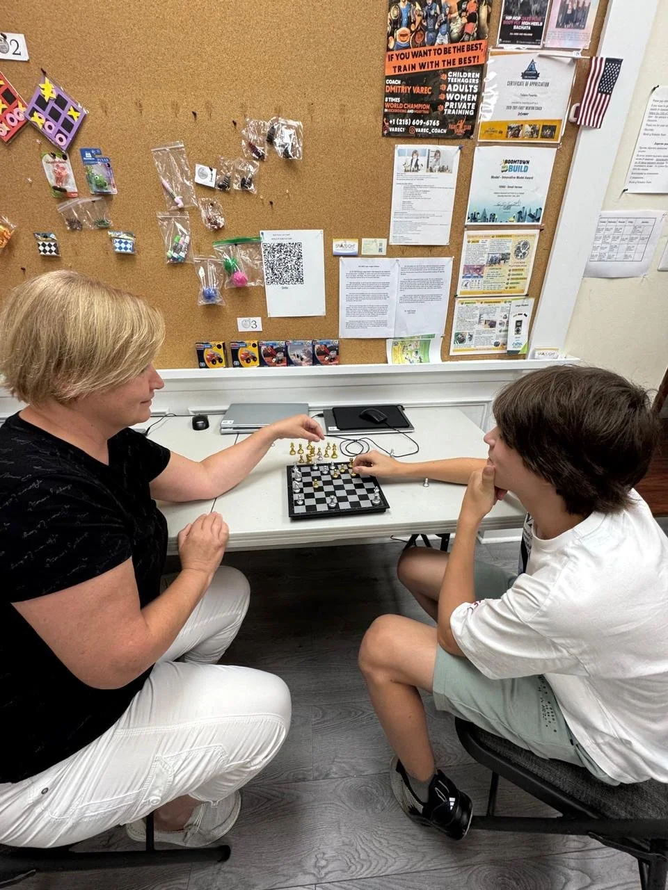 A woman and a teenager playing chess at a small table in a room with a brown corkboard filled with posters, papers, and small items hanging on the wall.