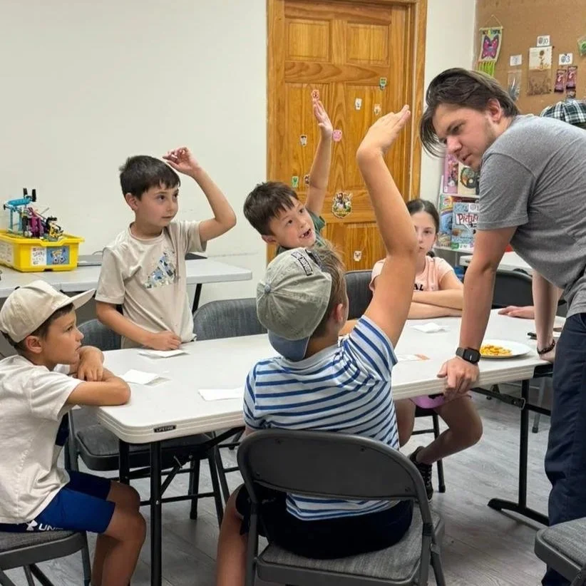 A man is leaning over a table to talk to children, with several kids raising their hands in a classroom setting.