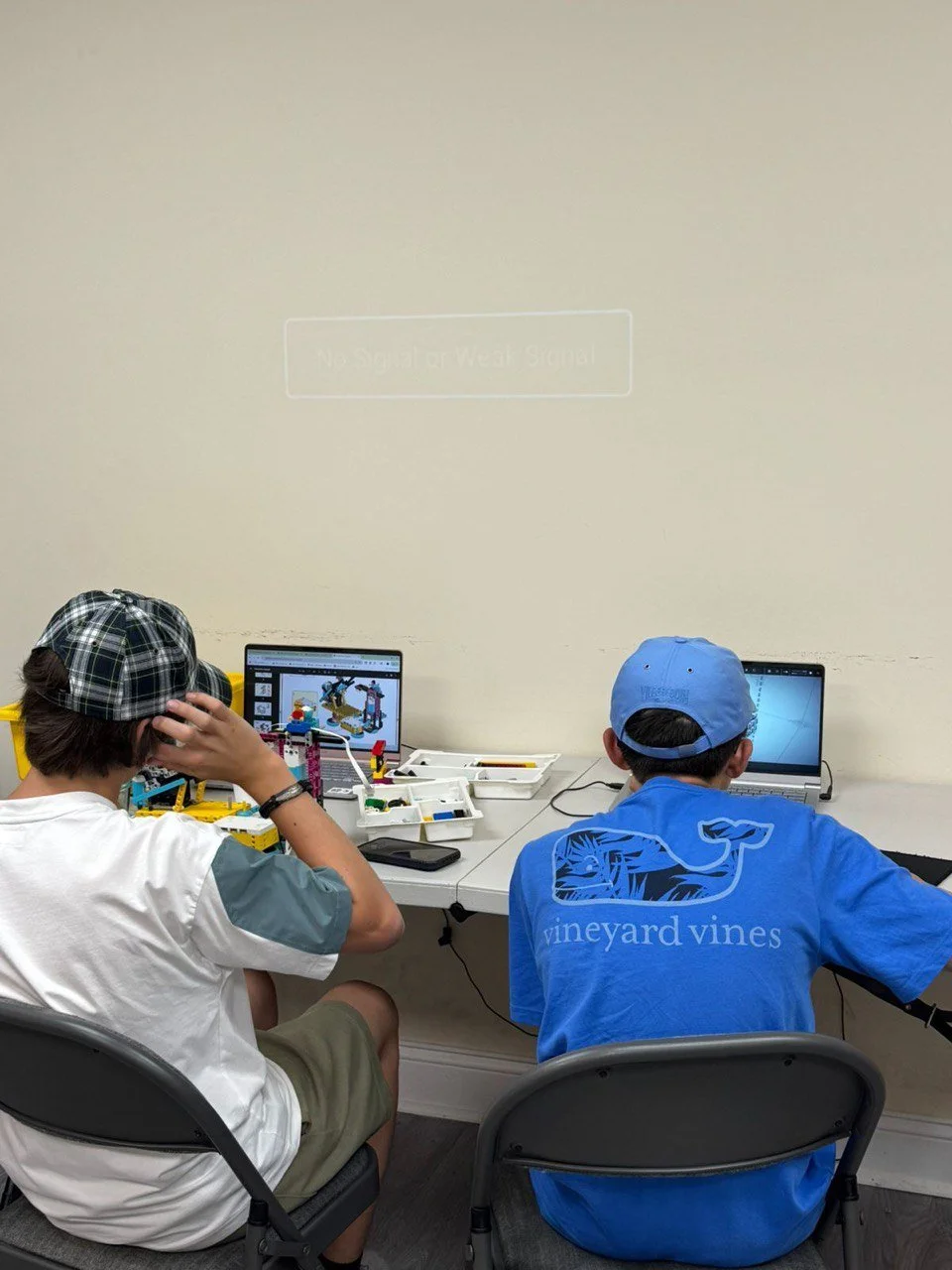 Two boys sitting at a table using laptops, with LEGO sets and materials in front of them, engaged in a building activity during a classroom or workshop.
