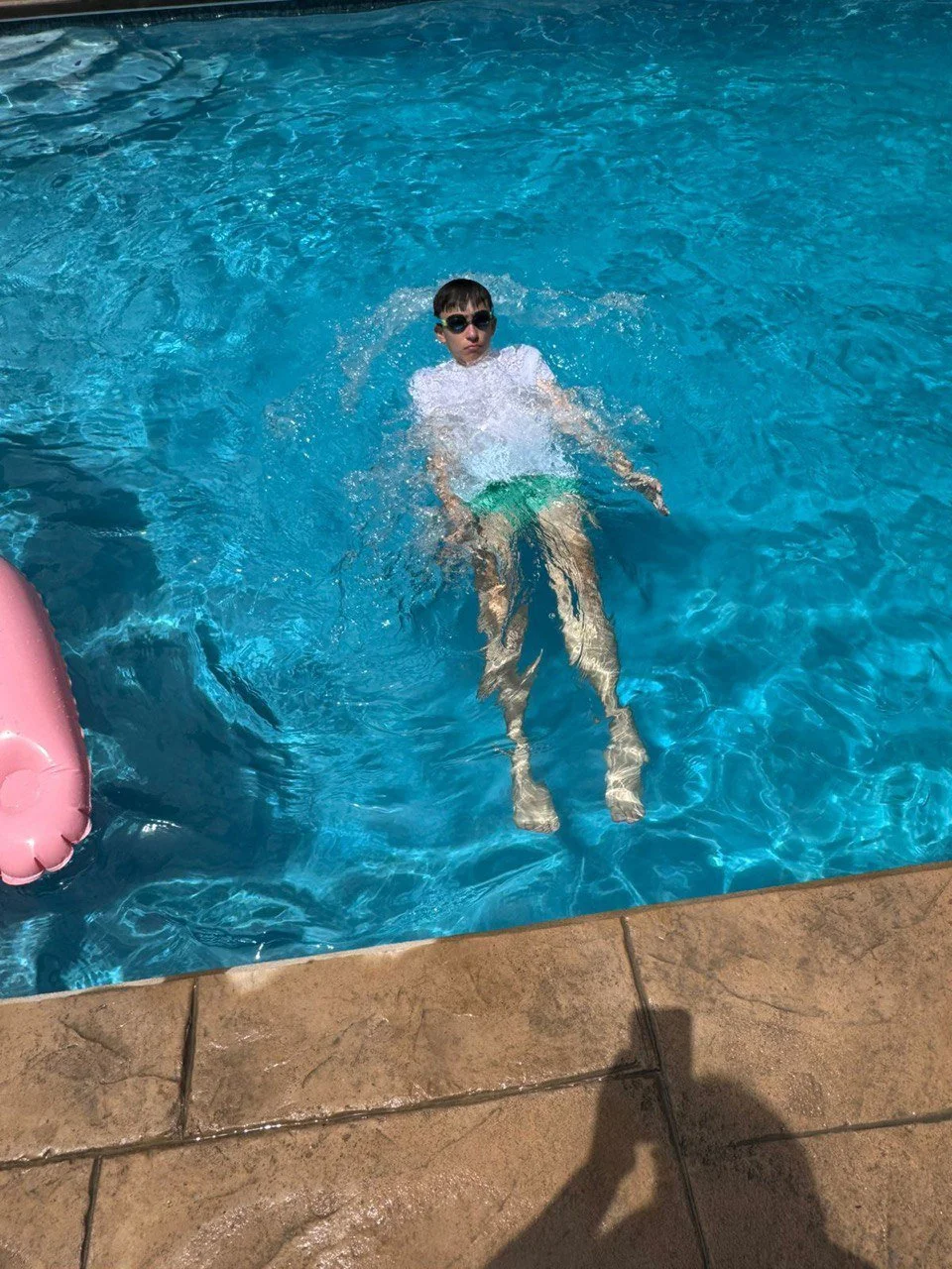 A boy wearing sunglasses, a white t-shirt, and green swim trunks floating in a swimming pool.