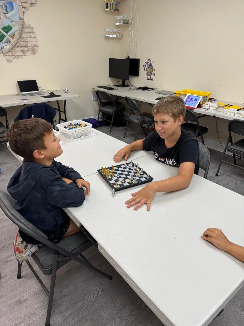 Two boys playing chess at a white table in a room with computers and shelves in the background.