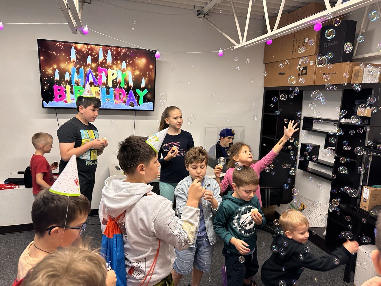 Children celebrating birthday party with birthday hats, blowing bubbles, and a birthday cake on a screen that says 'Happy Birthday' with lit candles and colorful letters.