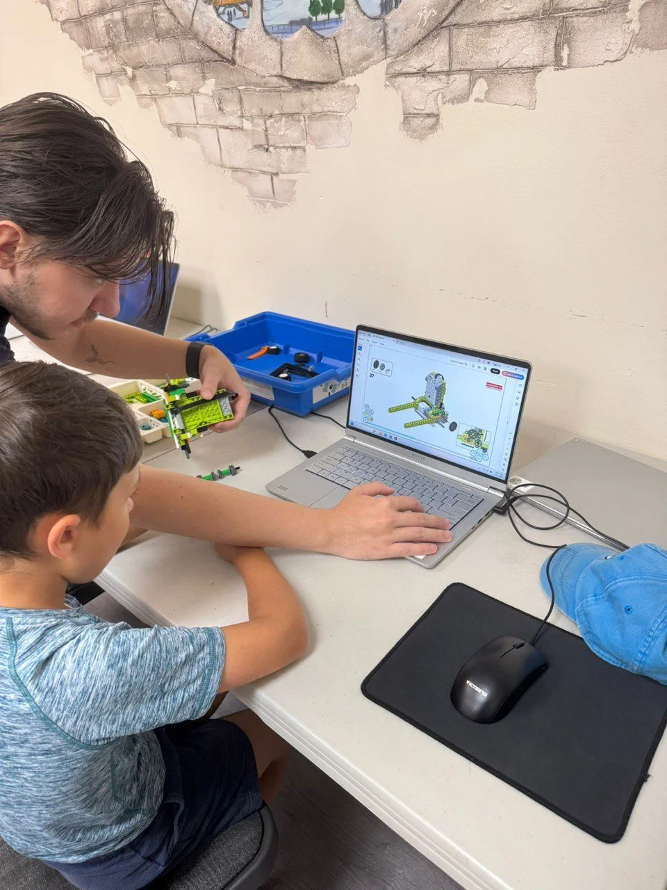 A man and a young boy are sitting at a desk with a laptop, computer mouse, and Lego robotics set. The man is helping the boy assemble the Lego model, which is shown on the laptop screen as part of a robotics design program. The setting appears to be 