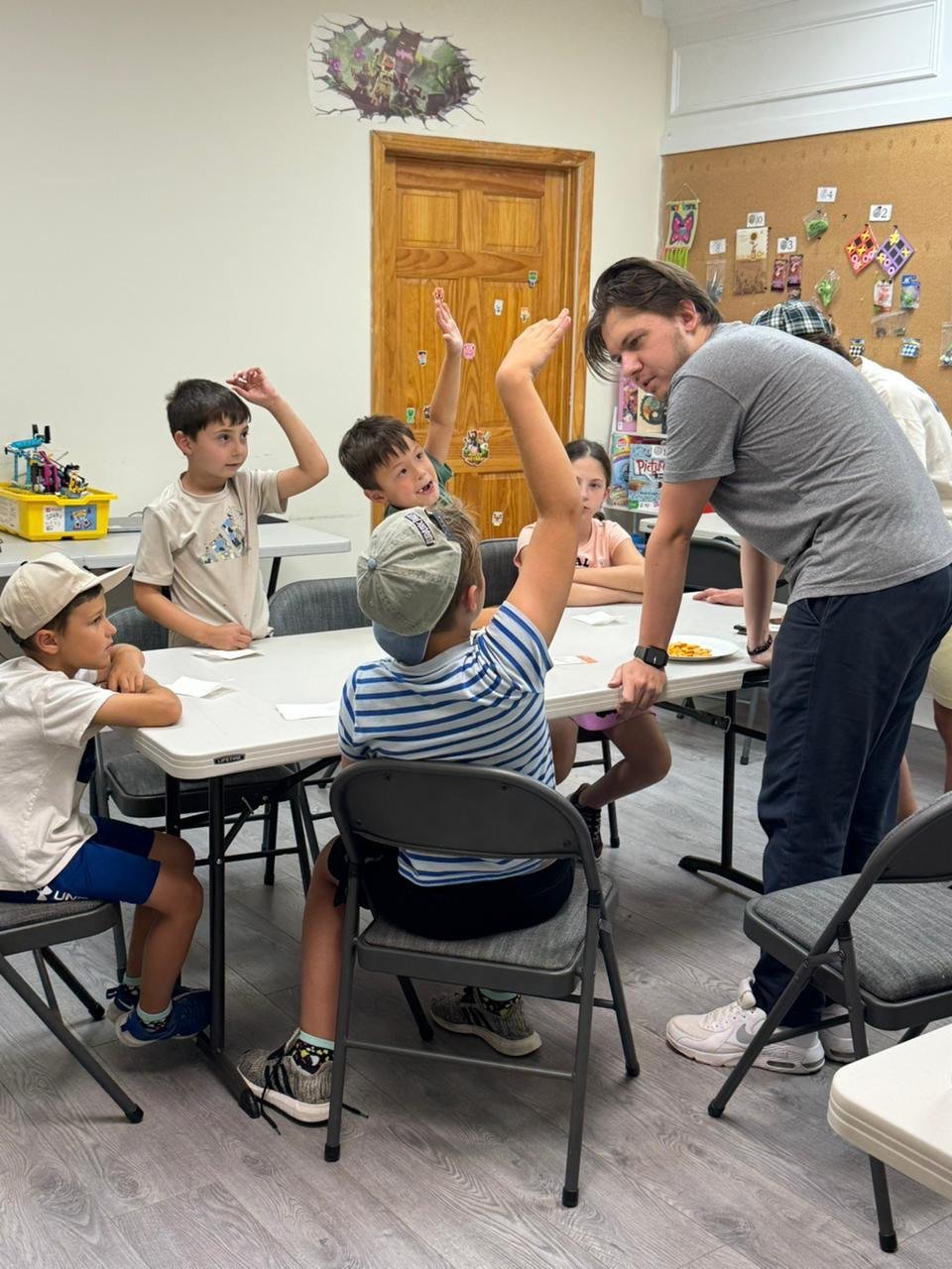 A group of children sitting at a table in a classroom, with one boy raising his hand. An adult man is leaning over the table, engaging with the children. There are colorful decorations and toys on the walls and table.