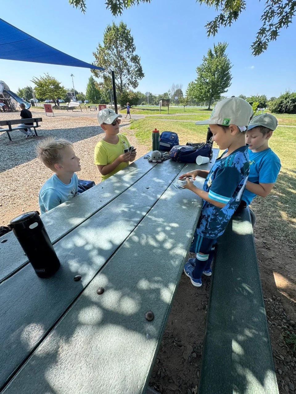 Four children are playing card games at a picnic table in a park on a sunny day, with trees and other park visitors in the background.