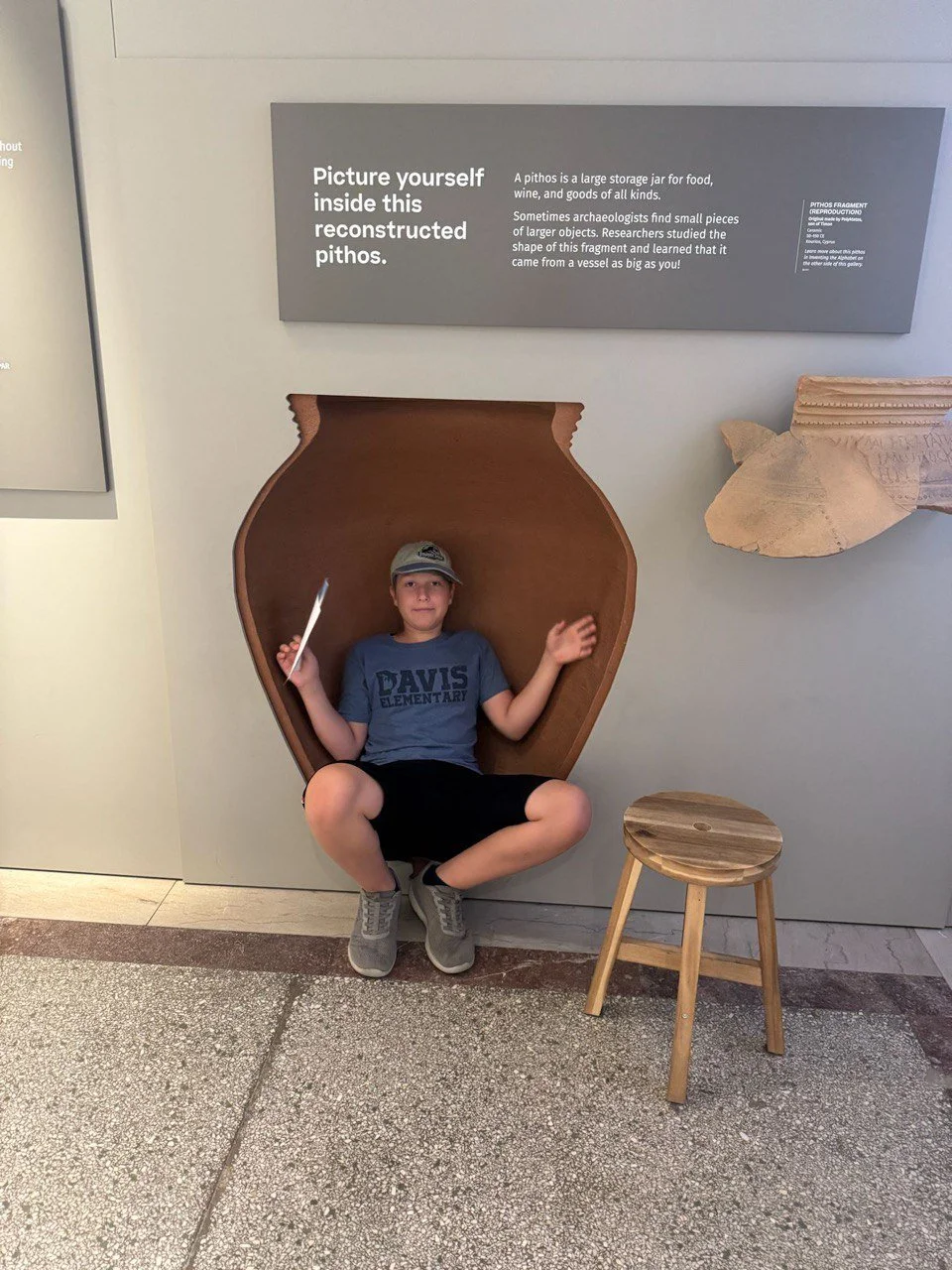 Child sitting inside a reconstructed pithos, a large storage jar, at a museum exhibit, holding a stick in one hand, with a small wooden stool next to him, and an informational sign above explaining the purpose of the pithos.