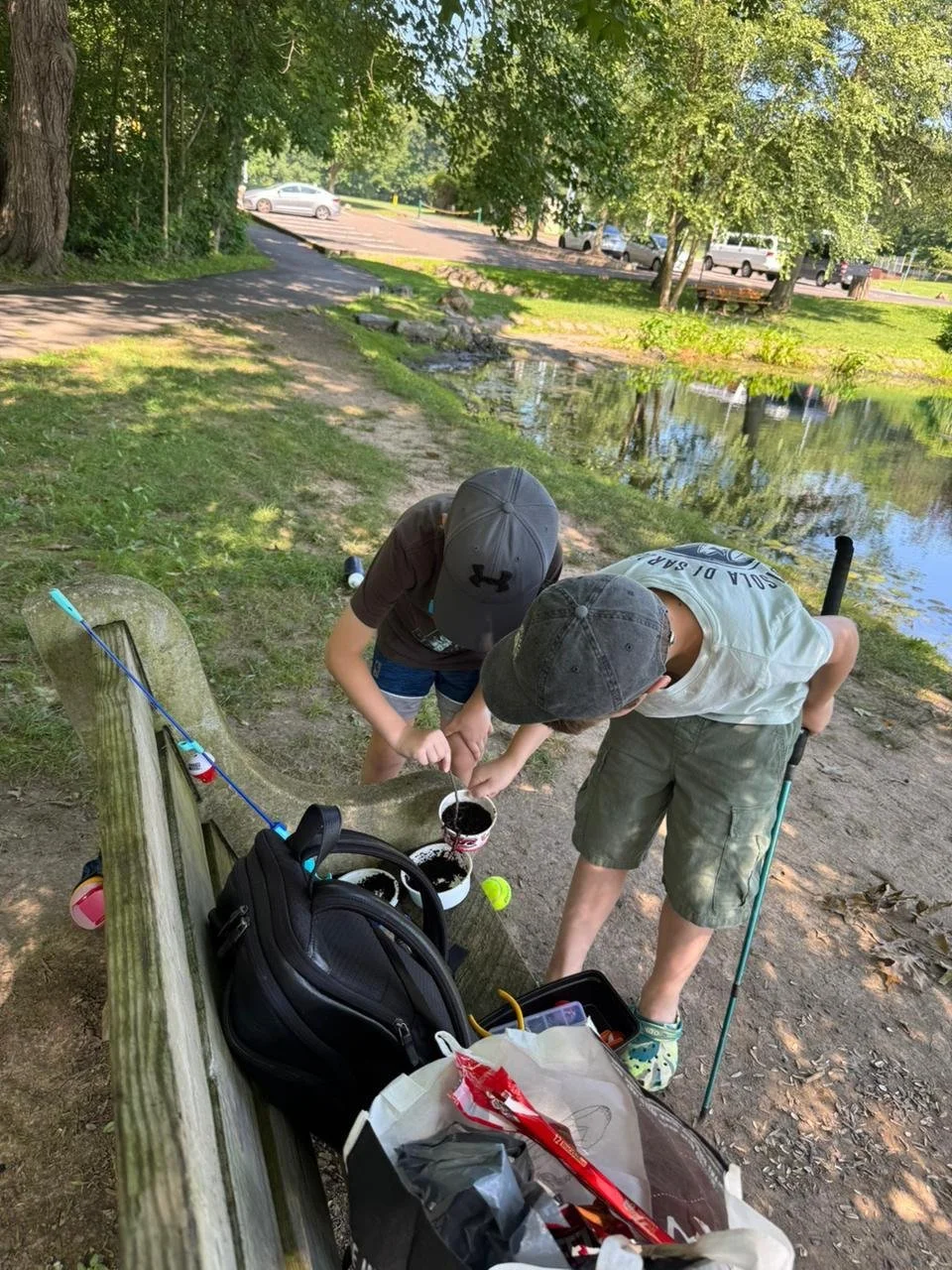 Two boys fishing by the water, standing next to a park bench with fishing gear and supplies around them.