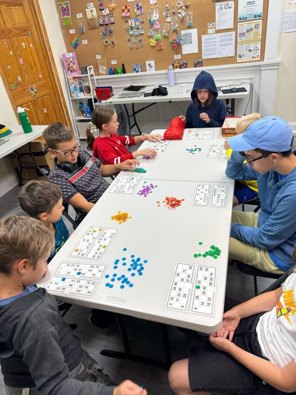 A group of children playing an educational game with colorful counters around a white table in a classroom.