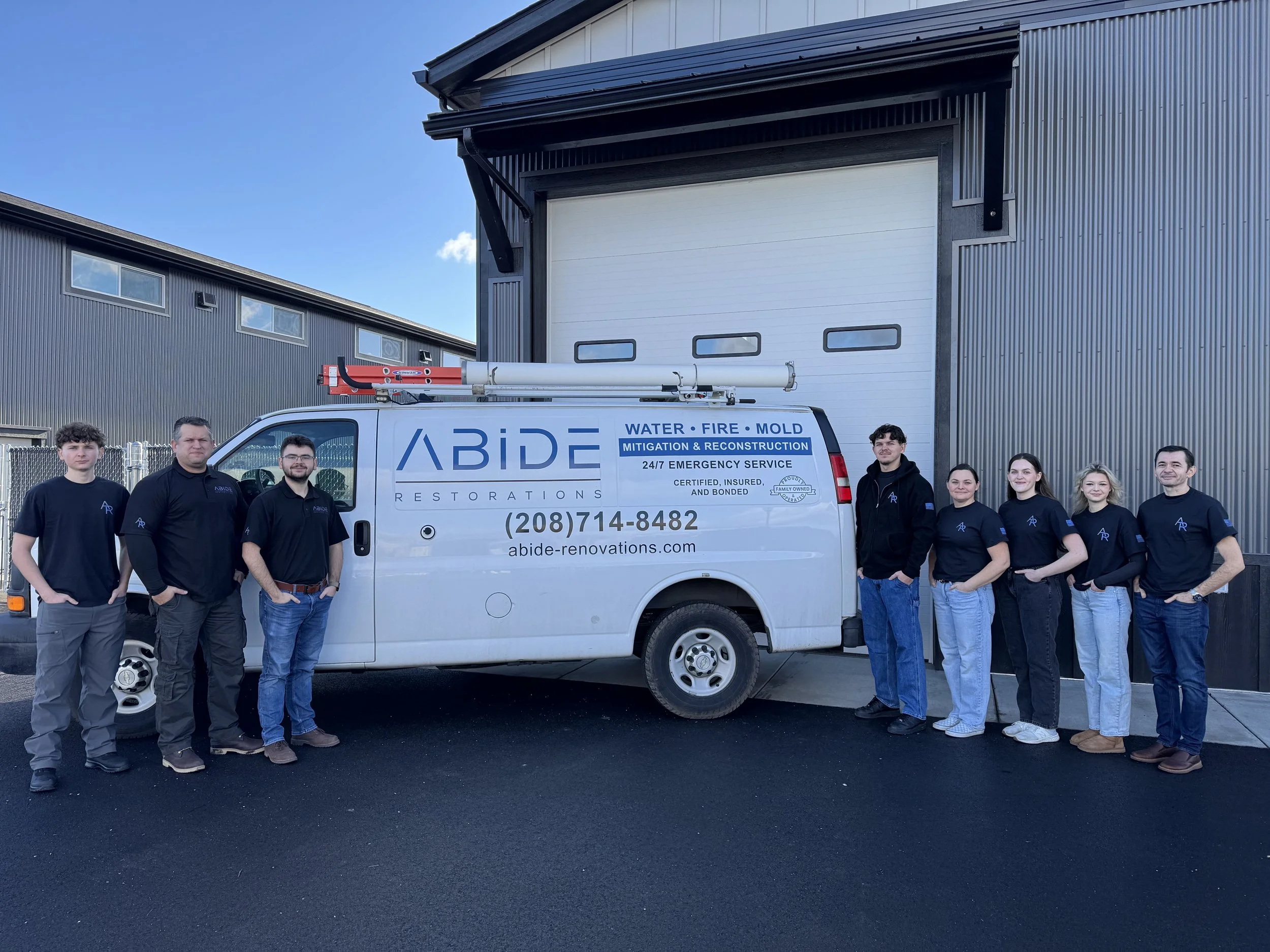 A team of nine workers standing in front of a white van with the company name 'Abide Restorations' printed on it. The van has equipment on top, and the team members are dressed in black and dark blue shirts, standing outside a building with large garage doors.