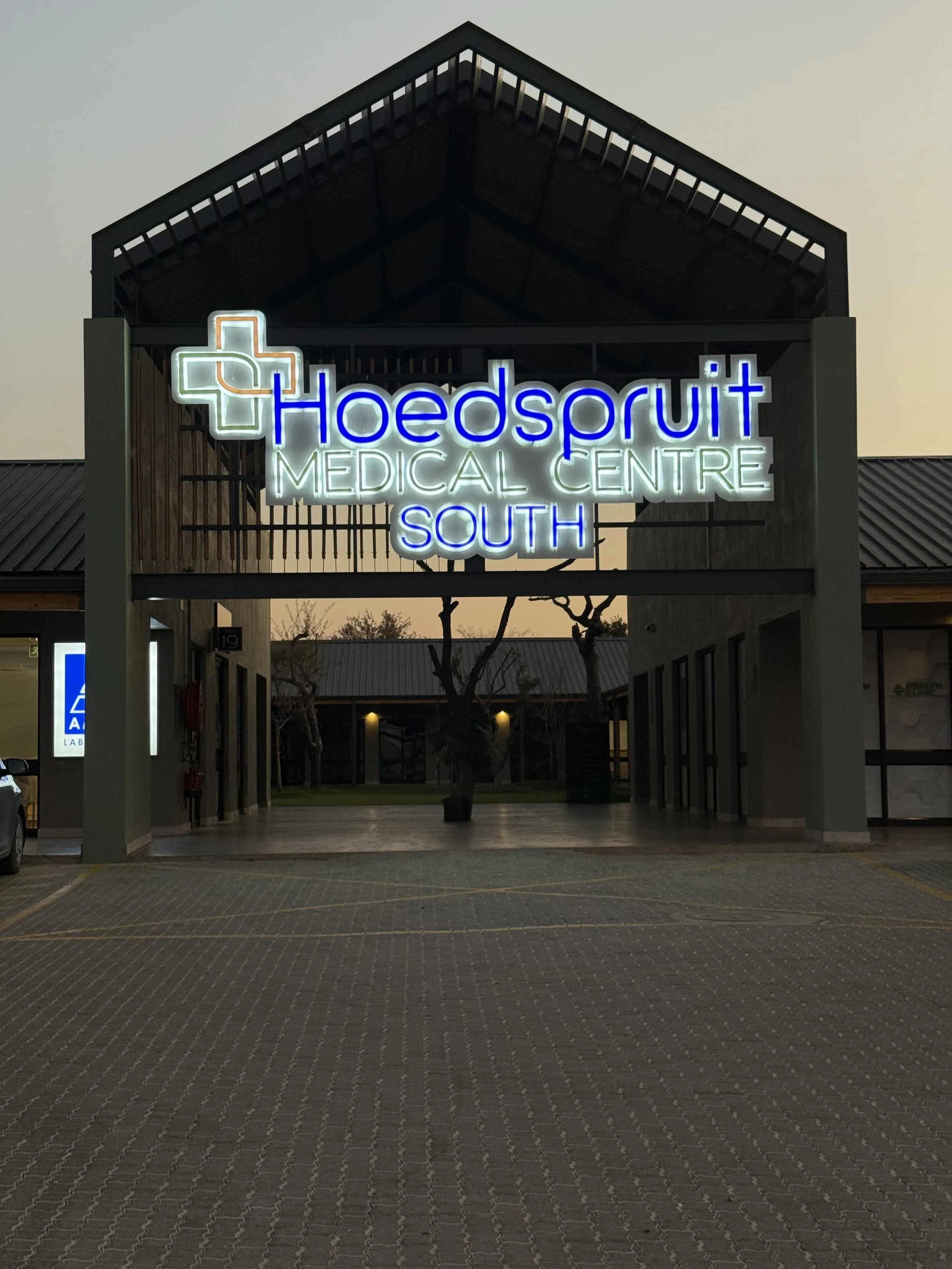 Exterior view of Hoedspruit Medical Centre South with a lit neon sign and a cross logo, evening setting with cloudy sky and empty parking lot.