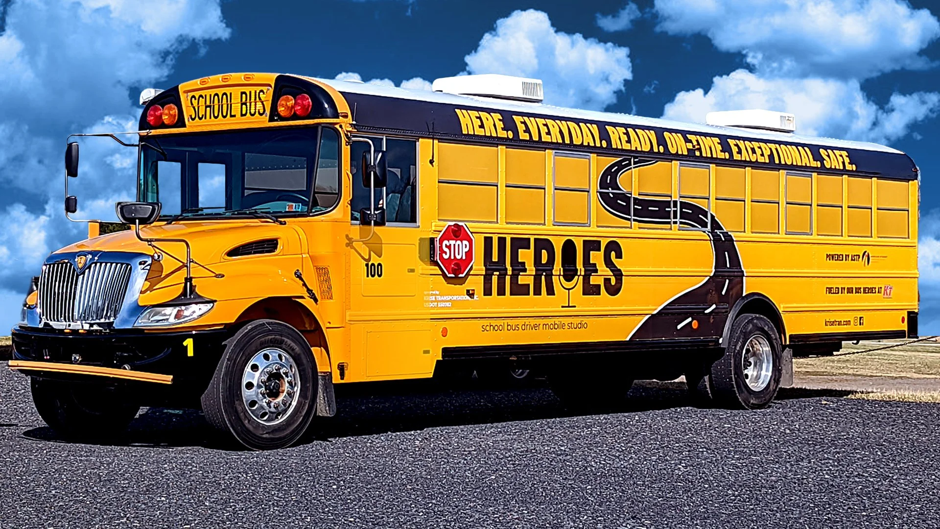 Yellow school bus with a road graphic and the word 'Heroes' on the side, parked on a road with a cloudy sky in the background.