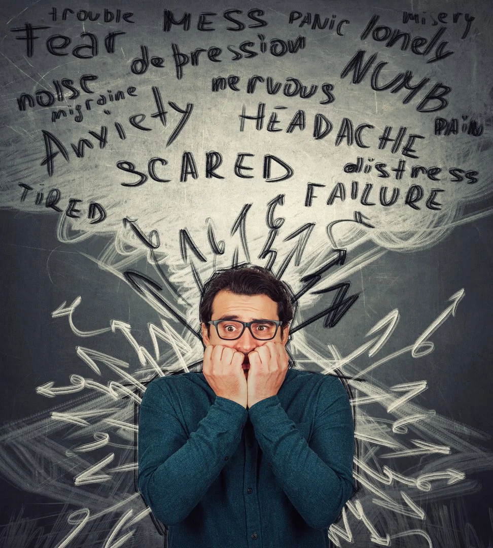 A person with glasses looking worried and covering their mouth in front of a chalkboard with words related to anxiety, fear, and mental health written around their head and shoulders, resembling an explosion or burst of thoughts.