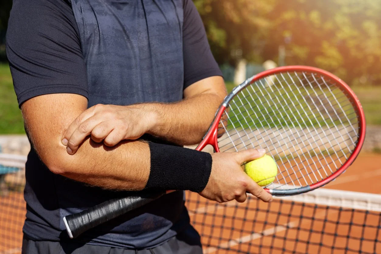 Person on the tennis court holding a tennis ball and racquet, wearing a black wristband, with a tennis net and trees in the background during sunset.