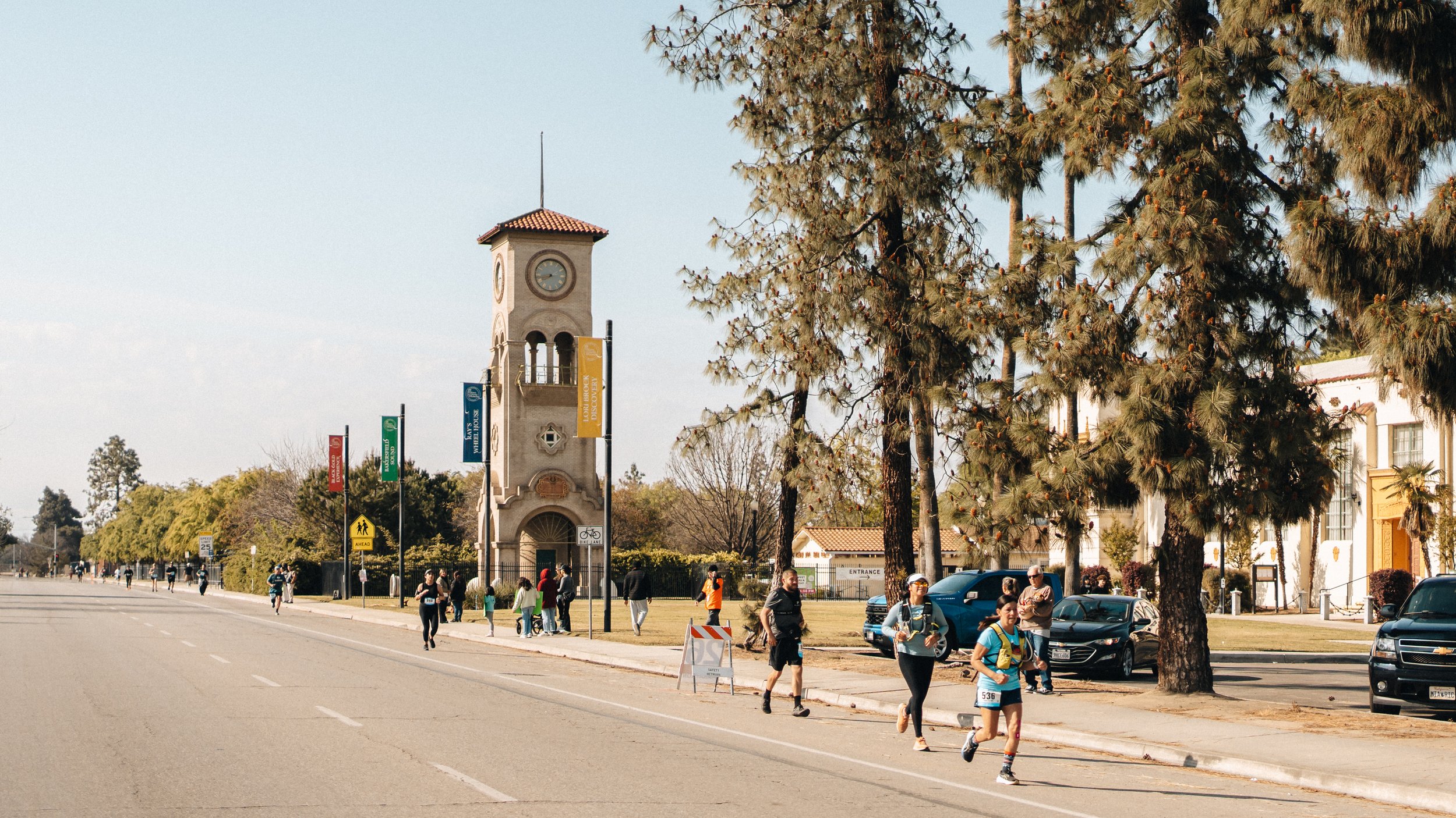 People participating in a road race or fun run along a city street lined with trees and cars, with a tall clock tower and colorful banners in the background.