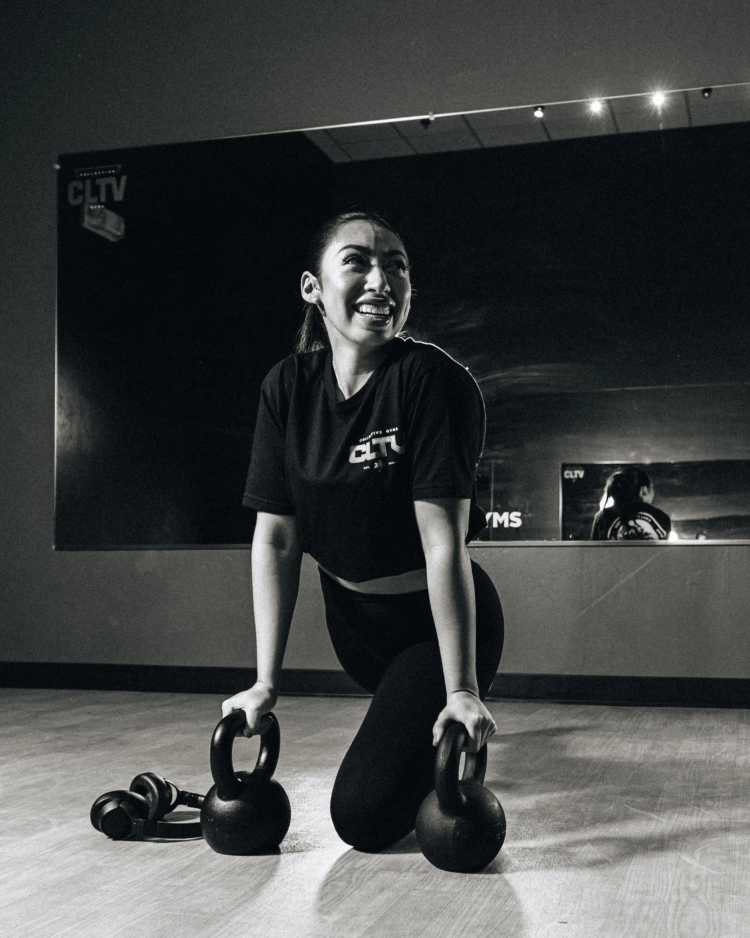A woman smiling while kneeling on the floor and holding kettlebells during a workout session at a gym.