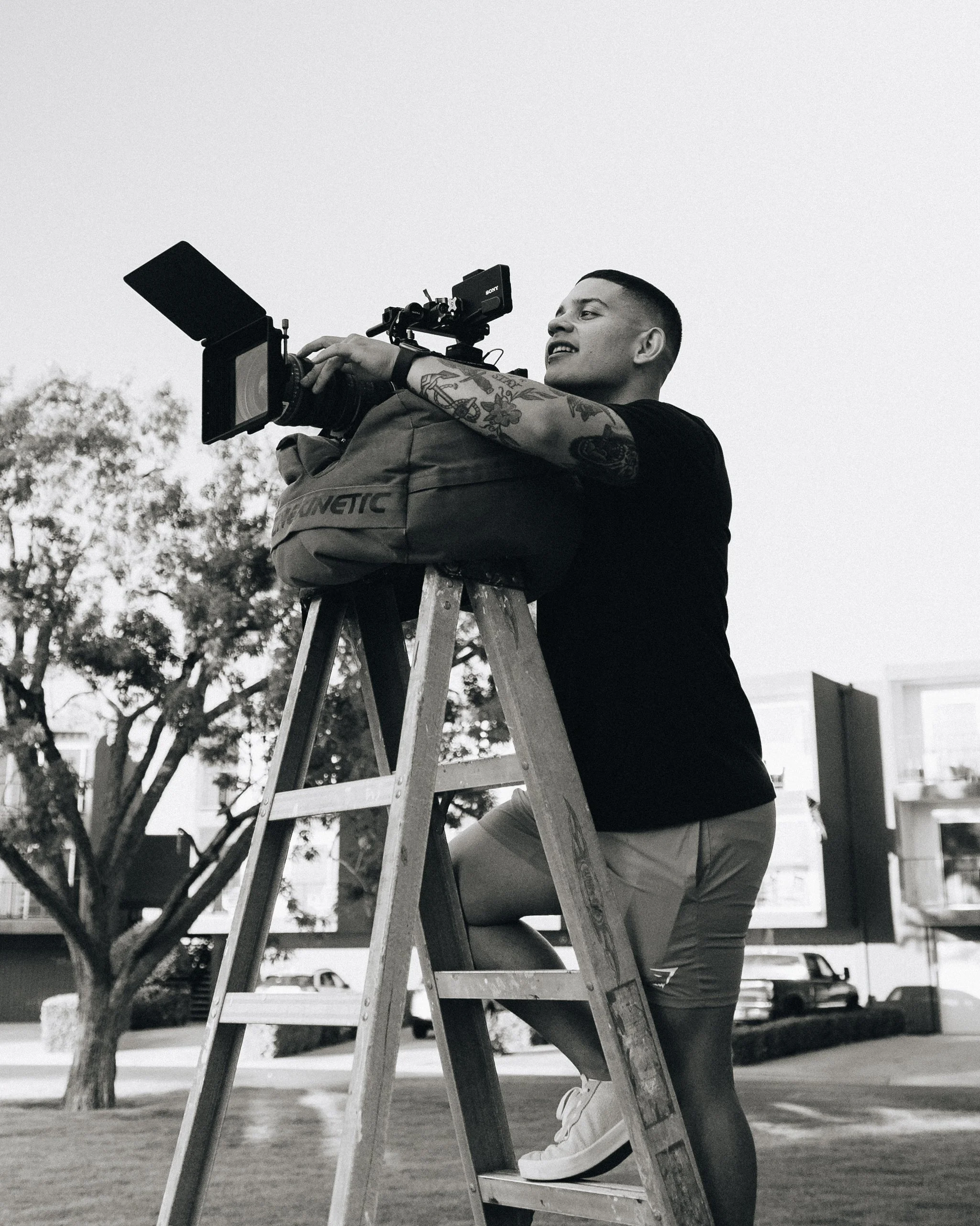 A person with tattoos filming with a professional camera on a ladder outdoors, with trees and buildings in the background.