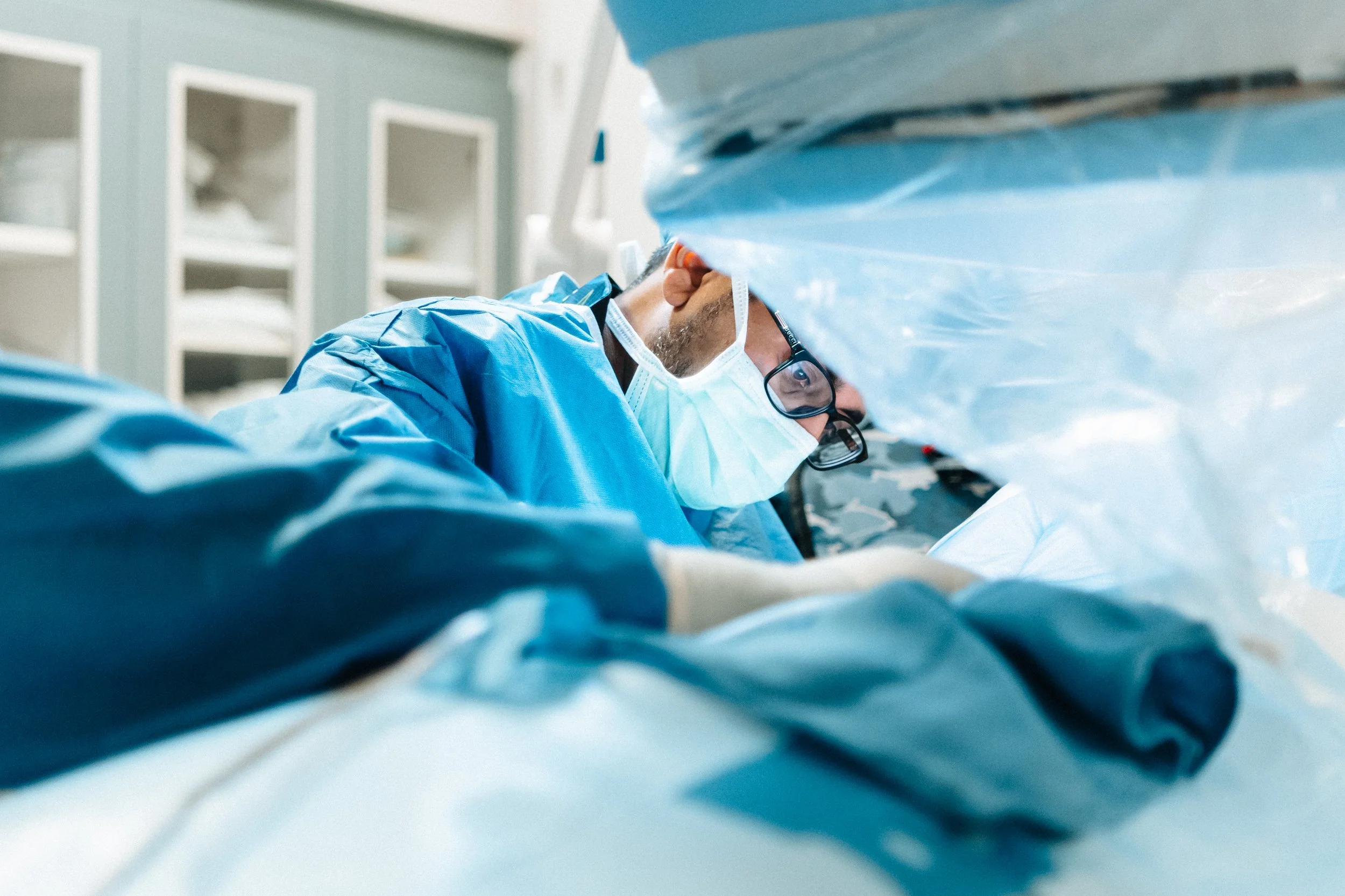 A surgeon wearing protective glasses, a face mask, and blue surgical gown is performing surgery in an operating room.