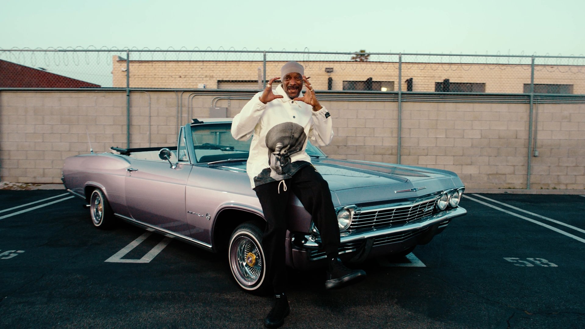 A man standing on a classic pink convertible car in a parking lot, gesturing with his hands.