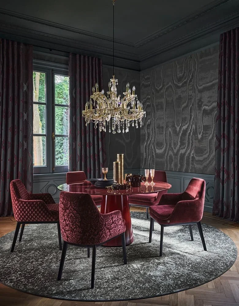 A luxurious dining room with a round red dining table, six velvet chairs in red tones, dark curtains, a chandelier, and a window showing greenery outside.