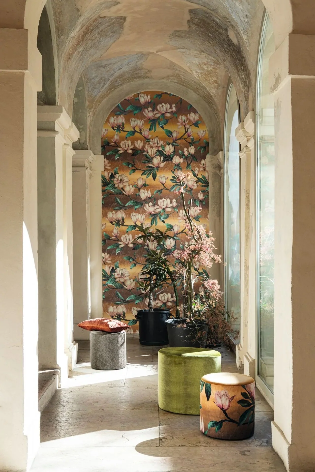 A bright interior space with high arched ceilings and large windows, decorated with floral wallpaper depicting blooming magnolia flowers and green foliage. Several potted plants and colorful stools are arranged along the space, with sunlight streaming through the windows.