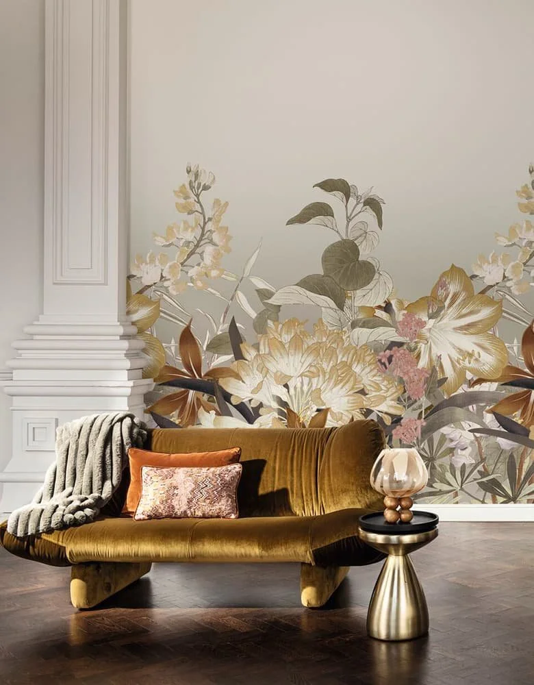 A vintage mustard-colored velvet sofa with two decorative pillows, one in pink and one in multicolored patterns. A gray knitted throw blanket is draped over the left arm of the sofa. Next to the sofa is a metallic side table with a gold finish, holding a beige lamp with a rounded glass shade and a base made of spherical shapes. The background features a wall with large floral wallpaper depicting flowers and leaves in soft pastel shades, with white decorative molding on the left side. The floor is dark hardwood.