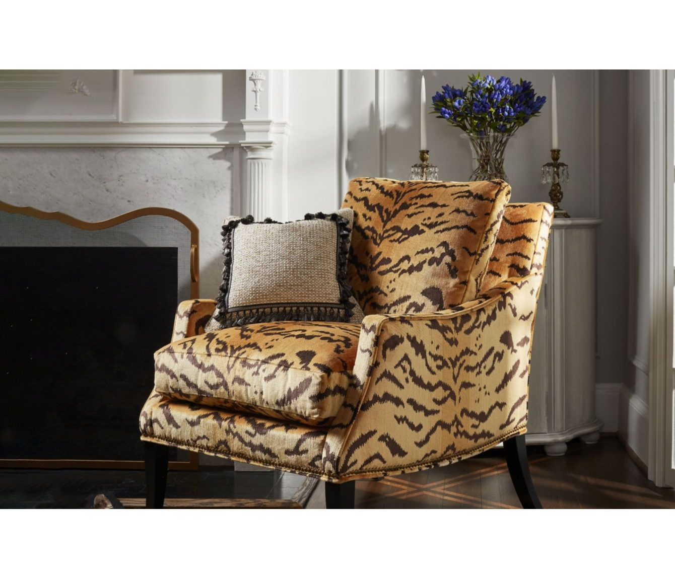 A leopard print upholstered armchair with beige and black accents, gray decorative pillow with black trim, situated in a room with white walls, a fireplace, a corner table with a blue bouquet, and tall white candles.