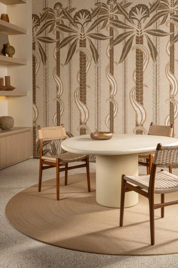 A round white table with three wooden chairs on a beige circular rug, with a decorative wallpaper featuring palm trees and plants in the background, and a cabinet with shelves and vases on the left.