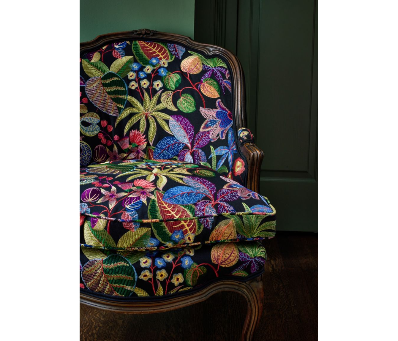 A vintage wooden armchair upholstered with a vibrant floral-patterned fabric against a dark green wall.