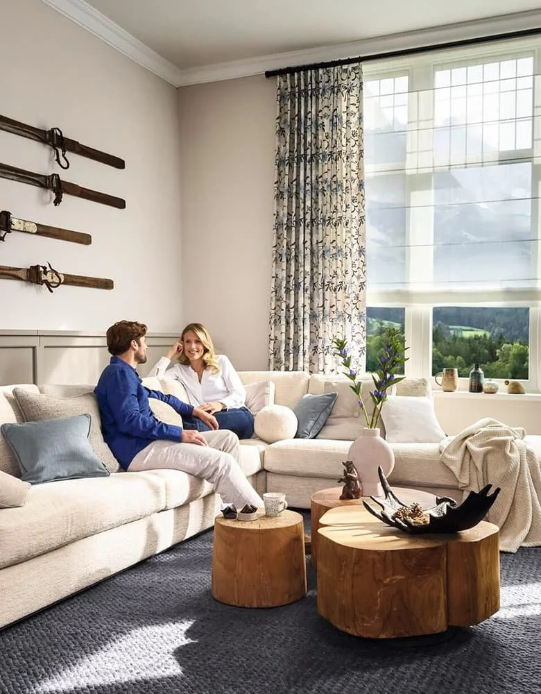 A cozy living room with a large window, cream-colored sectional sofa, two people sitting and chatting, wooden coffee tables, decorative vase with flowers, and wall-mounted swords.