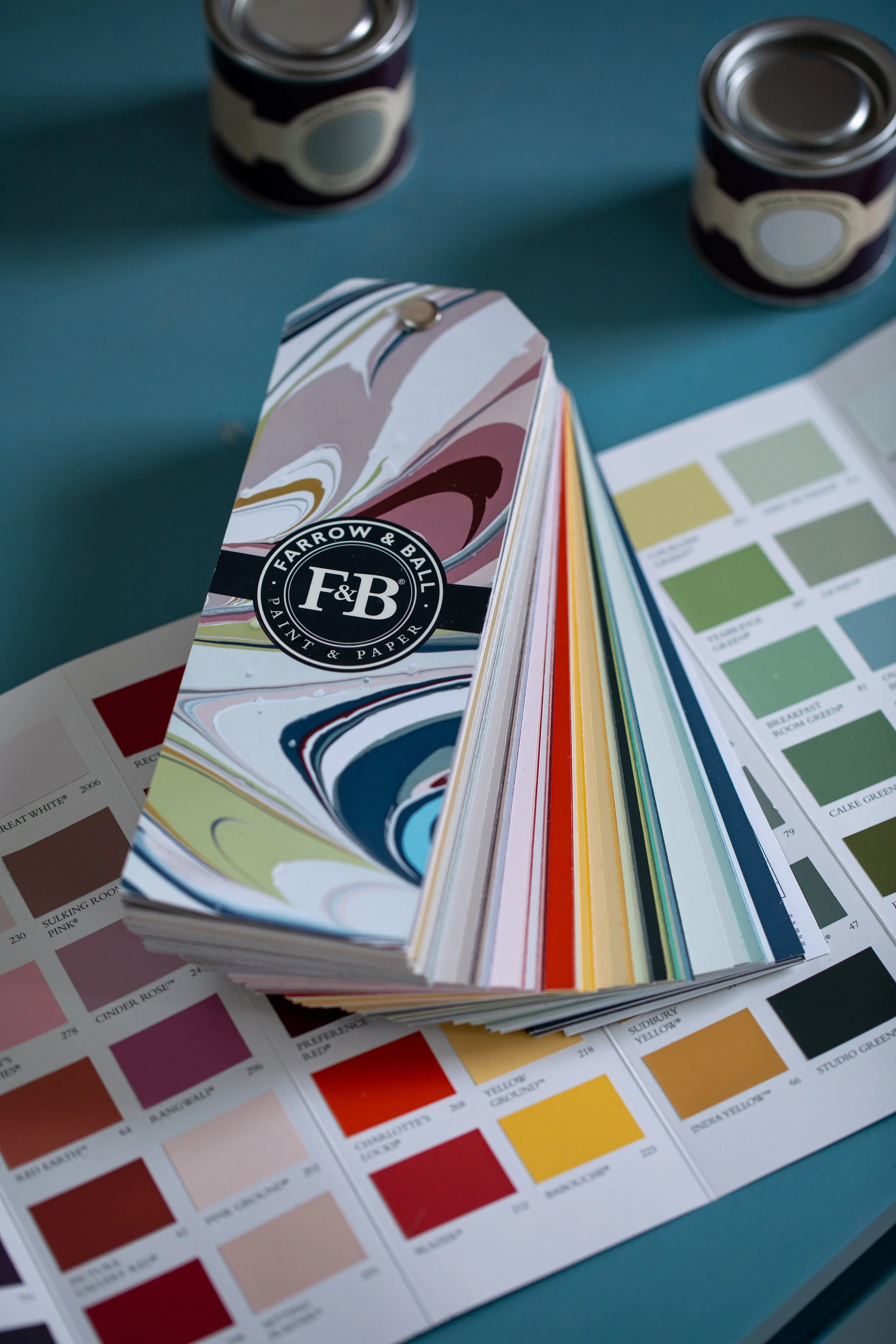 Color swatch book with paint color samples on a teal surface, with cans of paint and color guide pages in the background.