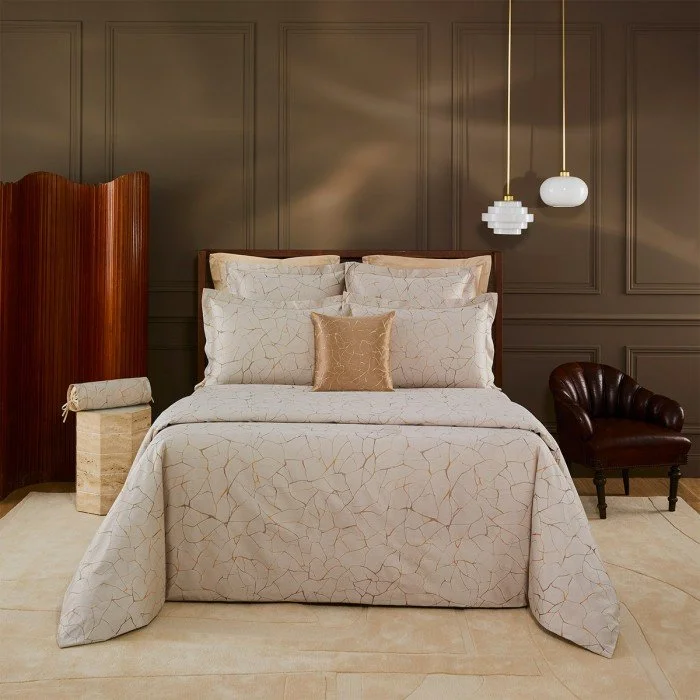 A neatly made bed with a white bedspread that has a subtle crackle pattern, and pillows including a smaller tan decorative pillow, set against a dark gray paneled wall. To the right, there's a small dark leather armchair, and to the left, a wooden room divider. Two modern pendant lights hang from the ceiling above the bed.