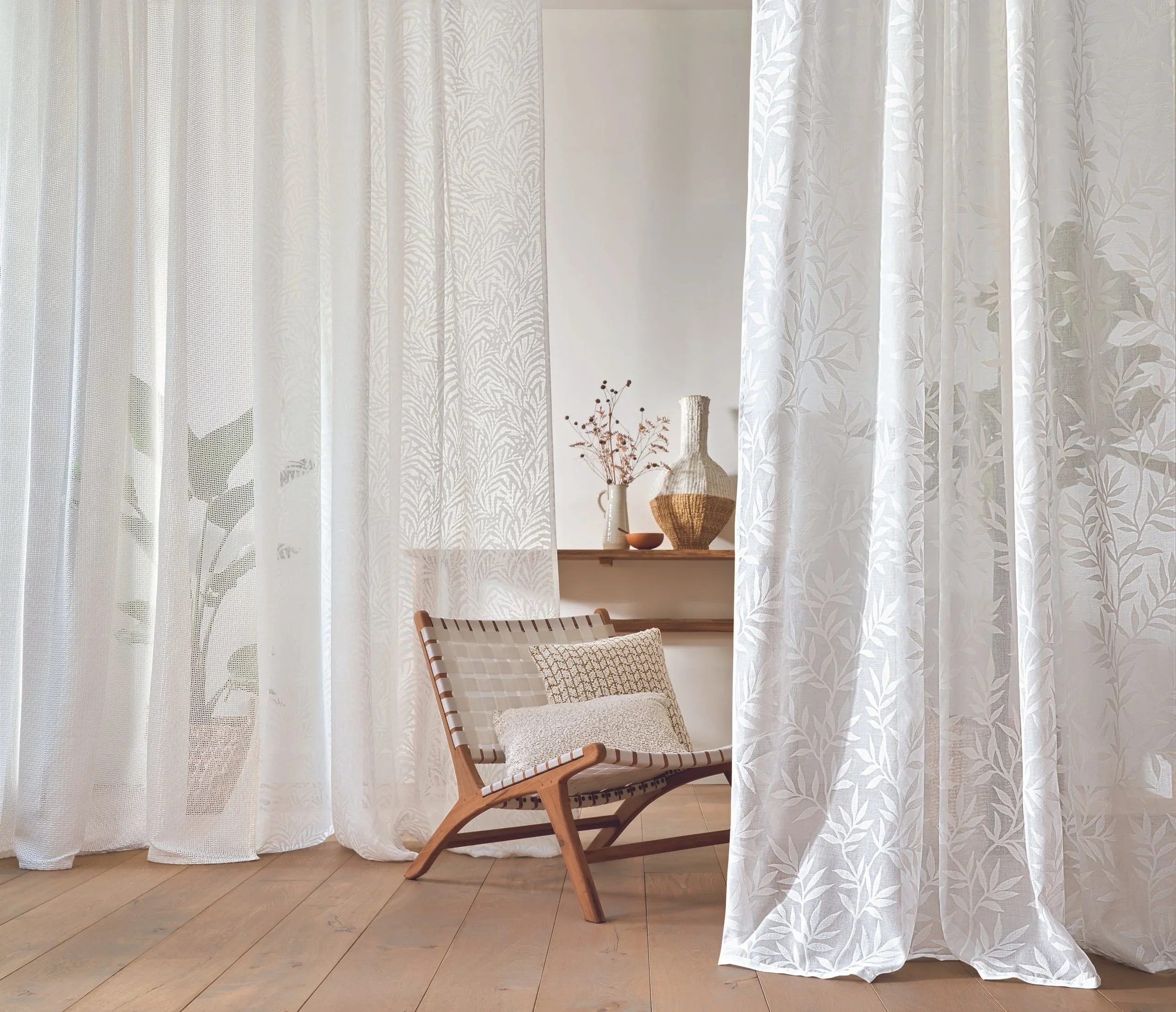 A cozy interior scene with sheer white curtains with leaf patterns, a wooden chair with woven fabric, and decorative vases with dried flowers on a wooden shelf.