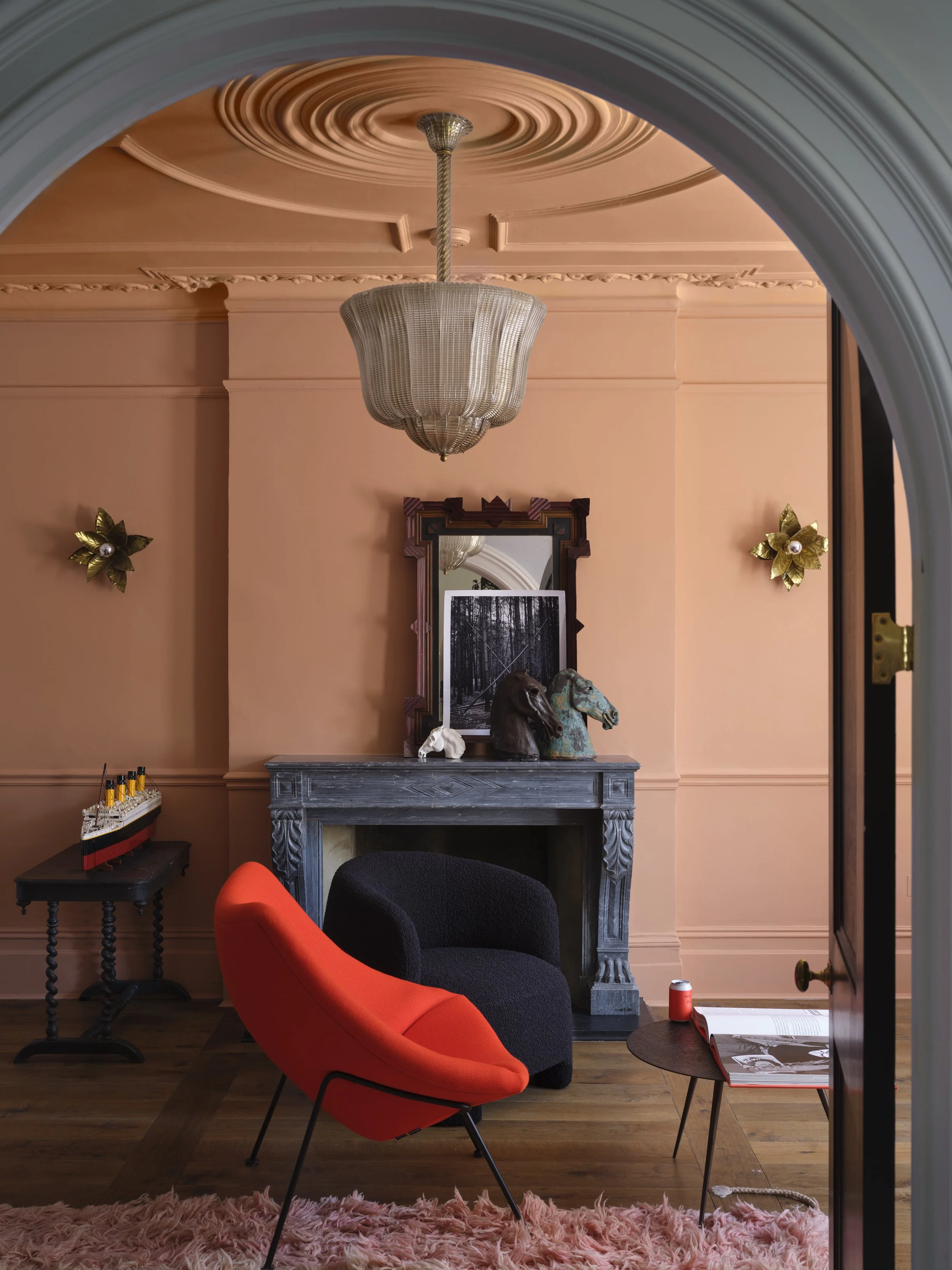Living room with salmon walls, a chandelier, a gray marble fireplace, two horse busts, a mirror, a red and black armchair, a small side table with books, a pink shag rug, and a gold flower wall decor.