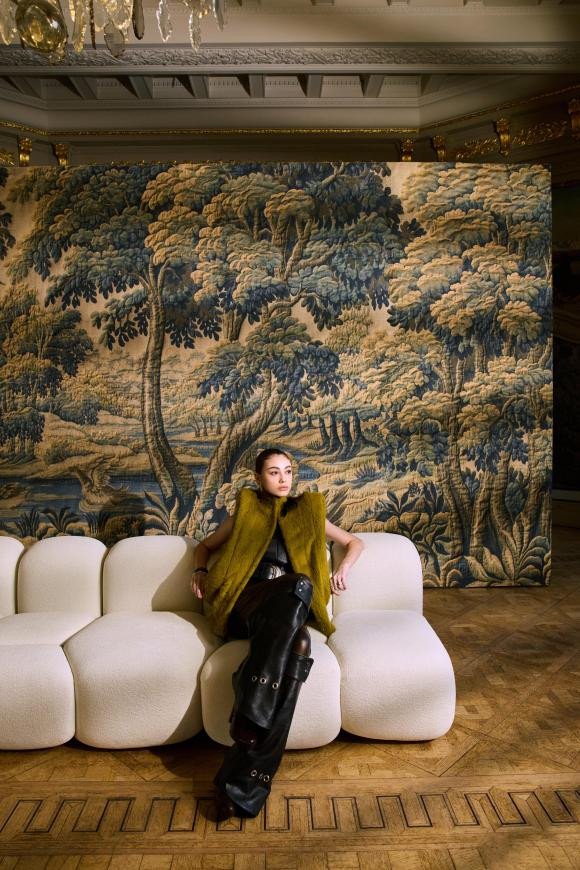 A woman in a green vest and black outfit sitting on a white, modern, plush couch in front of a large tapestry depicting a forest scene with tall trees and dense foliage. The setting appears to be an elegant, historic interior with decorative molding and a chandelier.