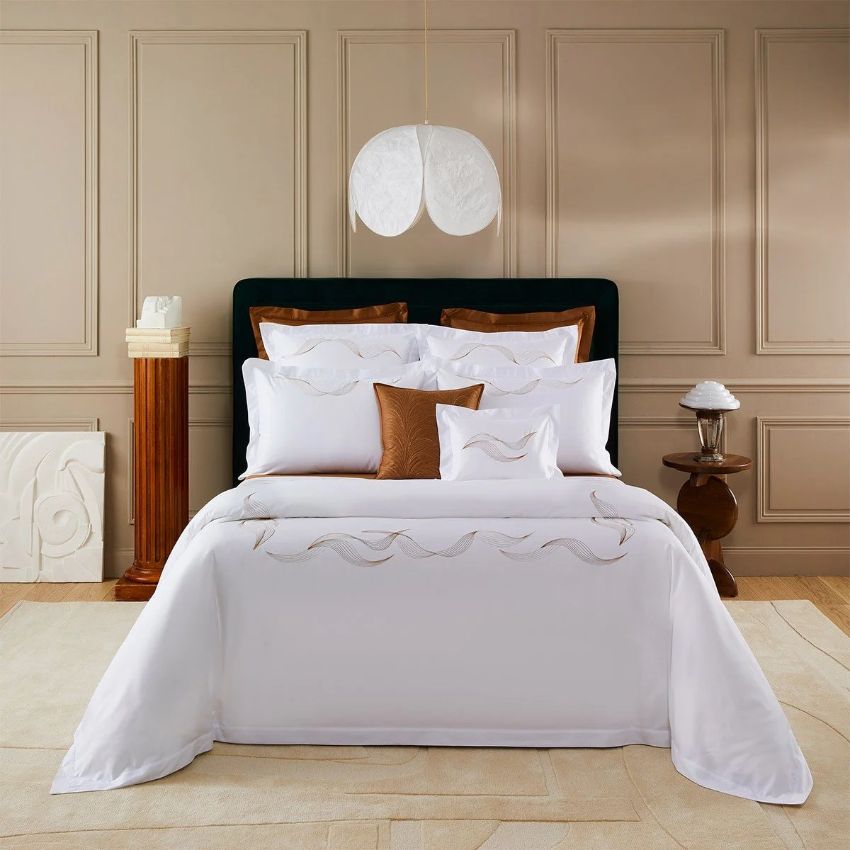 A neatly made bed with white and brown decorative pillows, black headboard, beige paneled wall, white pendant light hanging above, wooden side tables with lamps, and a cream-colored rug.