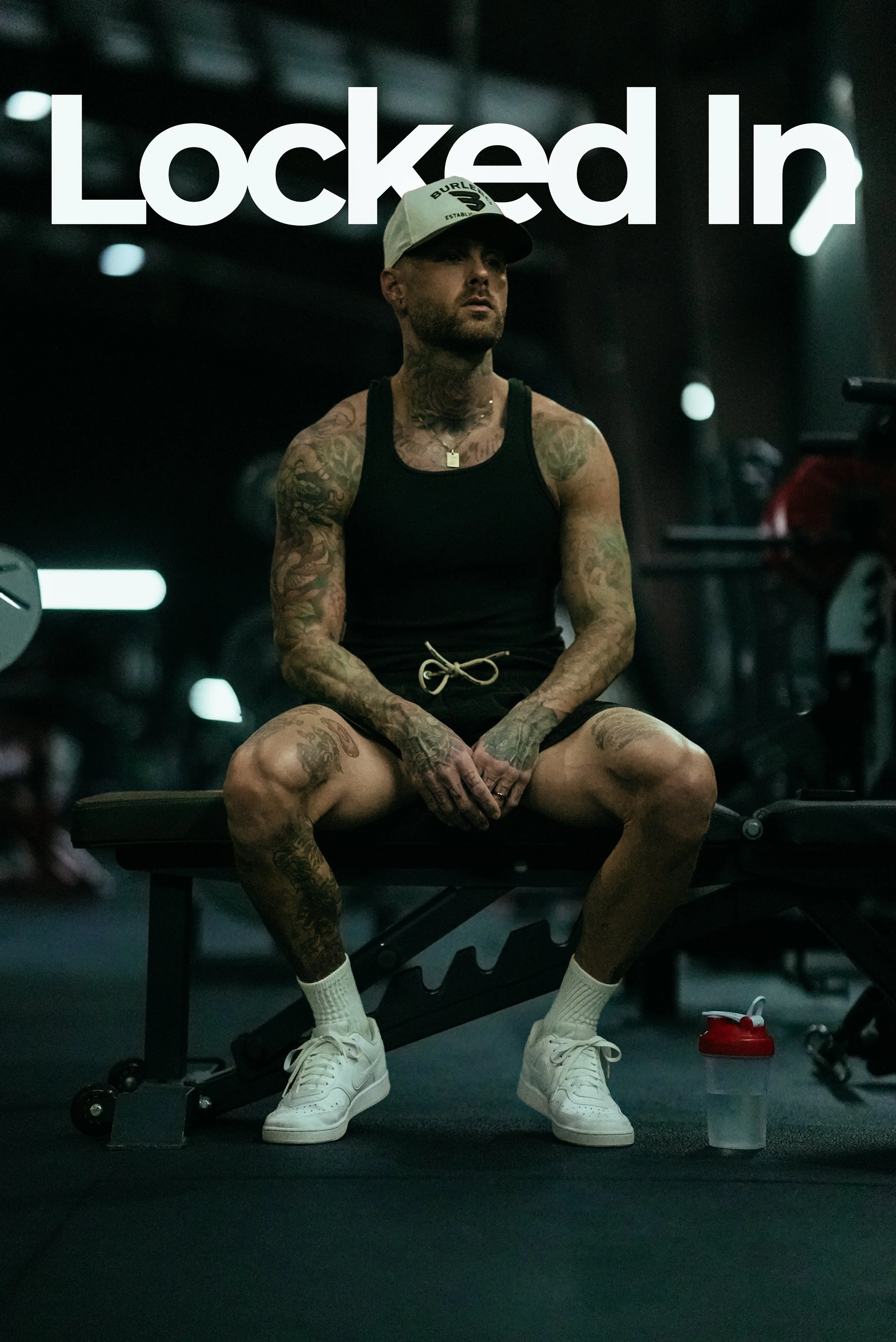 A tattooed man sitting on a workout bench in a gym with the words 'Locked In' displayed above him.