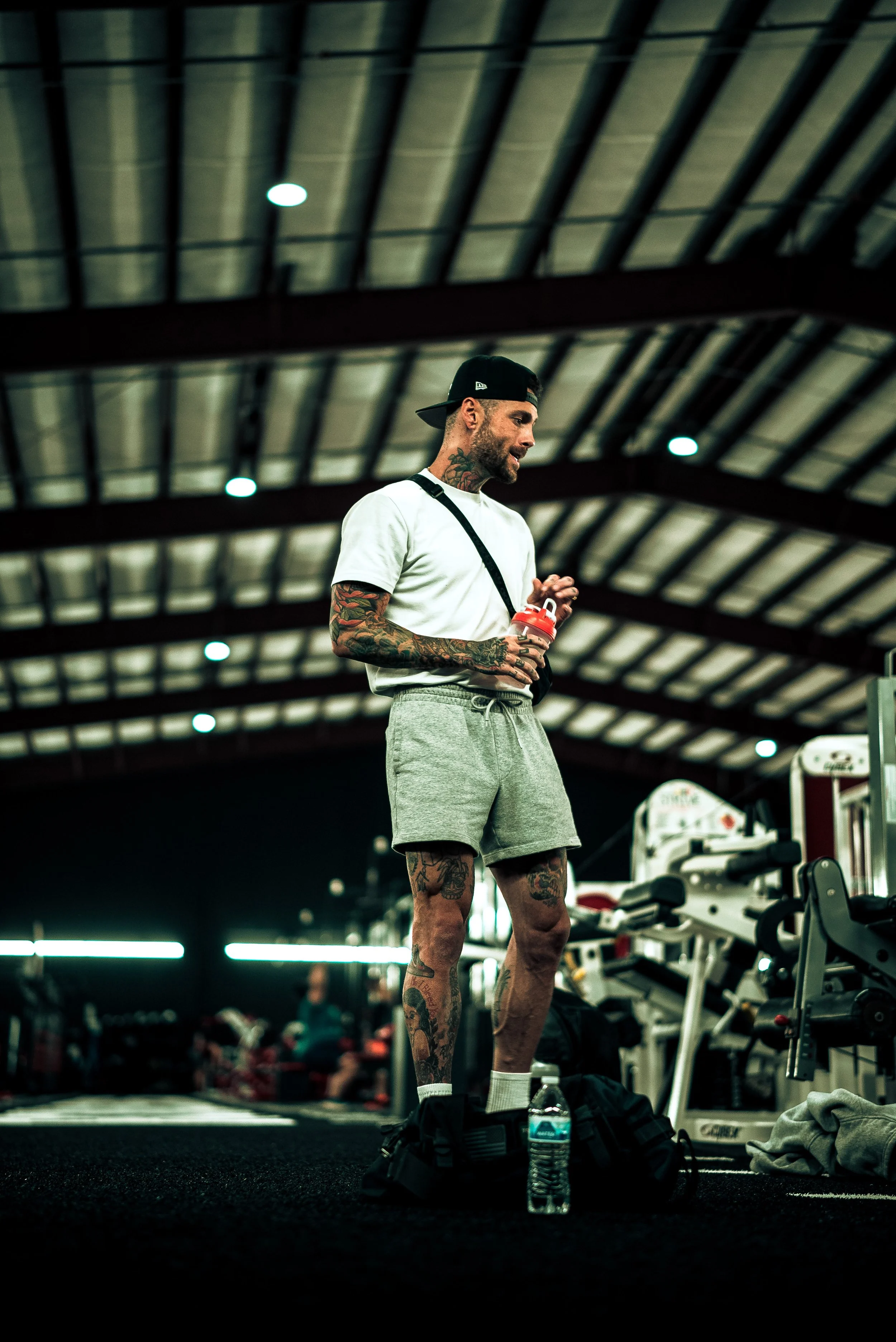 Tattooed man in a white t-shirt, grey shorts, and a backwards black cap standing in a gym, holding a water bottle.
