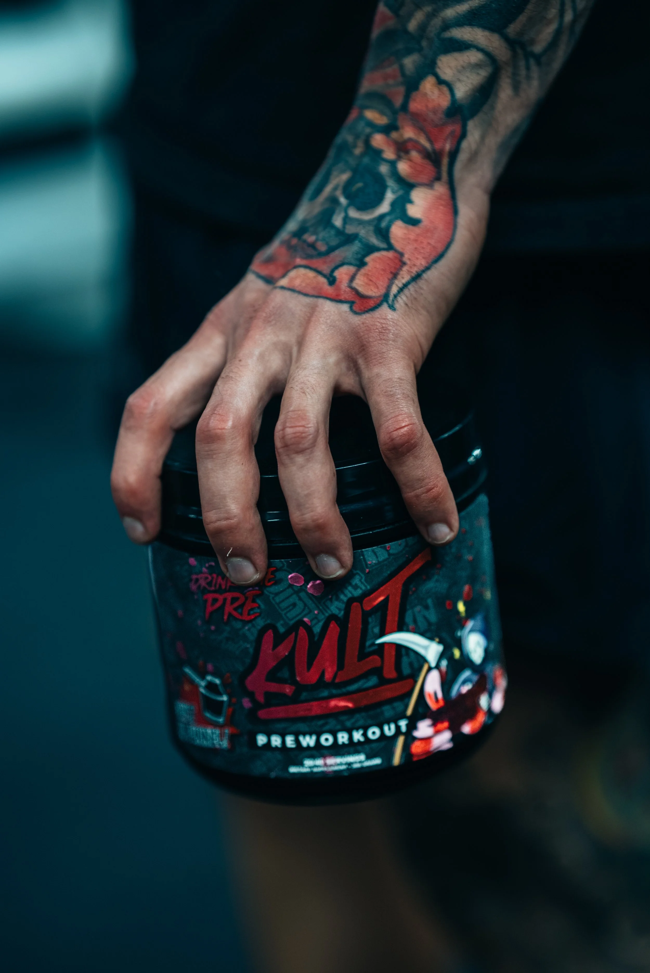 A person's tattooed hand gripping a container of pre-workout supplement called 'Kult' with a black lid, with the background blurred.