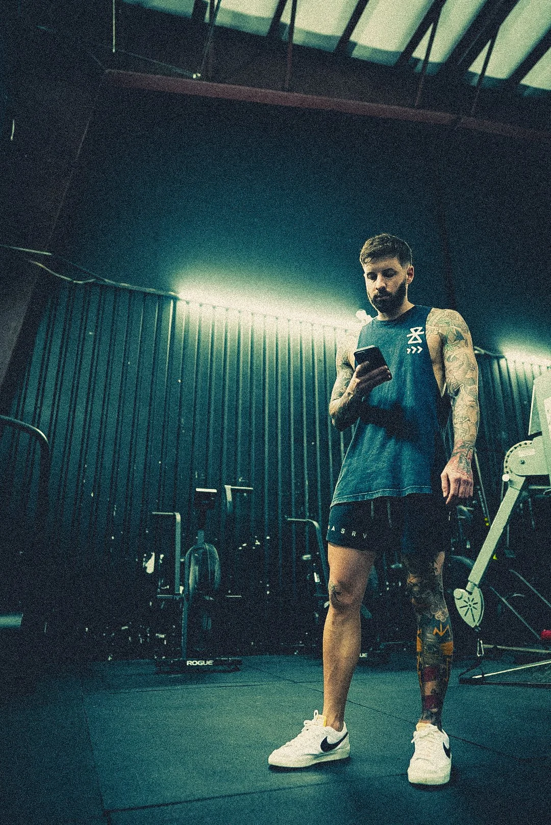 A man with a tattooed arm, wearing a athletic tank top and shorts, standing in a gym while looking at his phone.