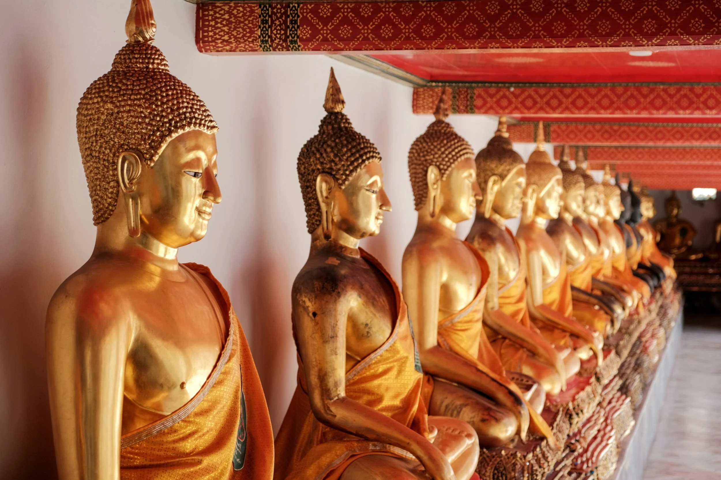 Series of golden Buddha statues sitting in a row, adorned in orange robes, inside a temple with red and gold decorative roof.