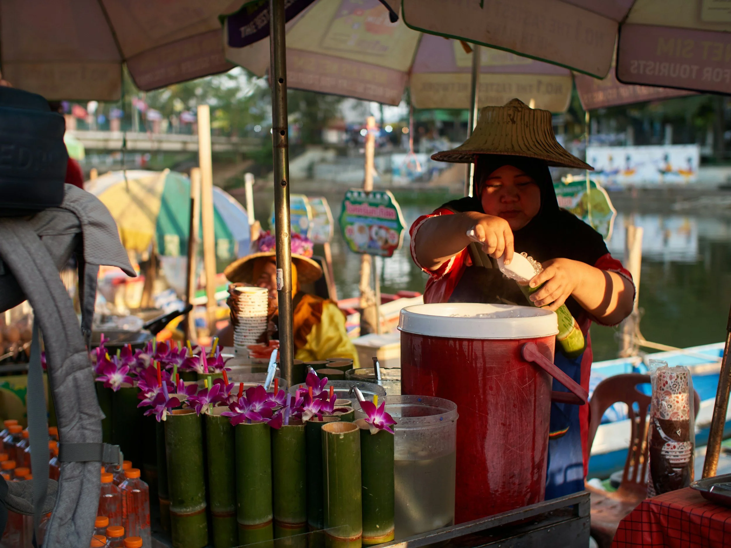 A woman wearing a conical hat prepares a beverage at a street food stall near a river, with decorative flowers in bamboo containers in the foreground.