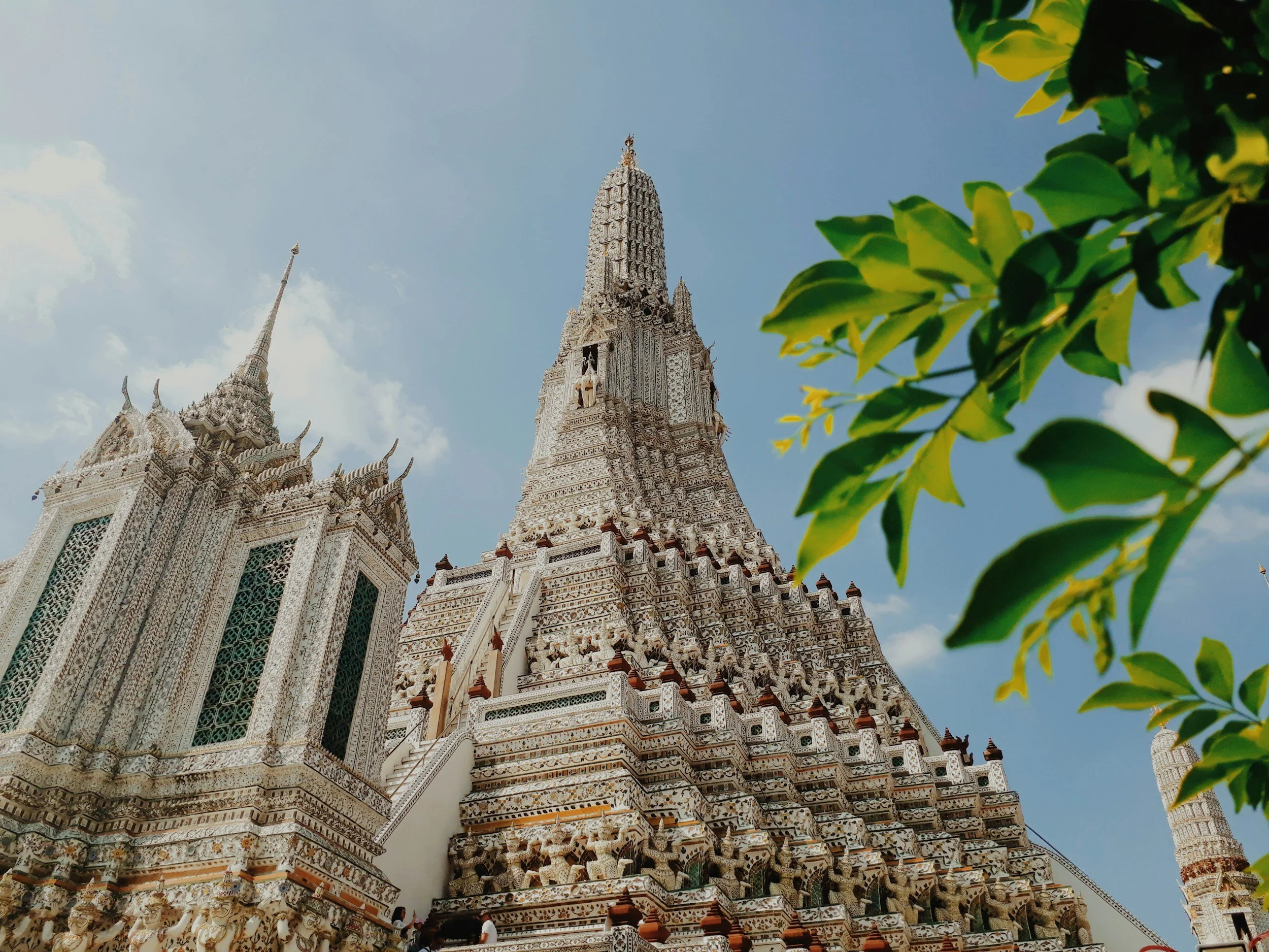 A detailed white temple with intricate carvings and multiple towers, set against a blue sky with some clouds, and green leaves partially visible on the right side.