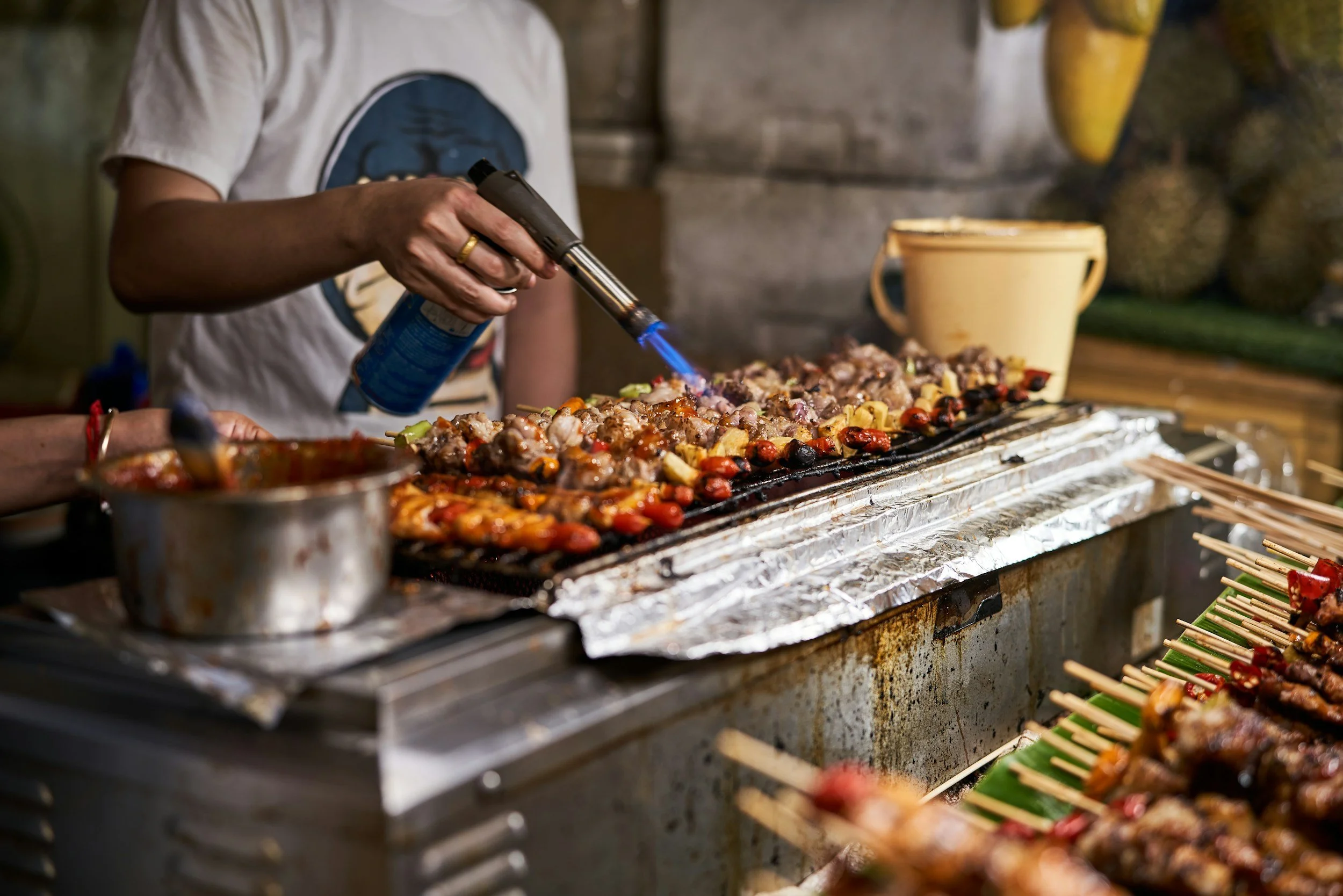 Person grilling skewers of meat and vegetables using a blowtorch.