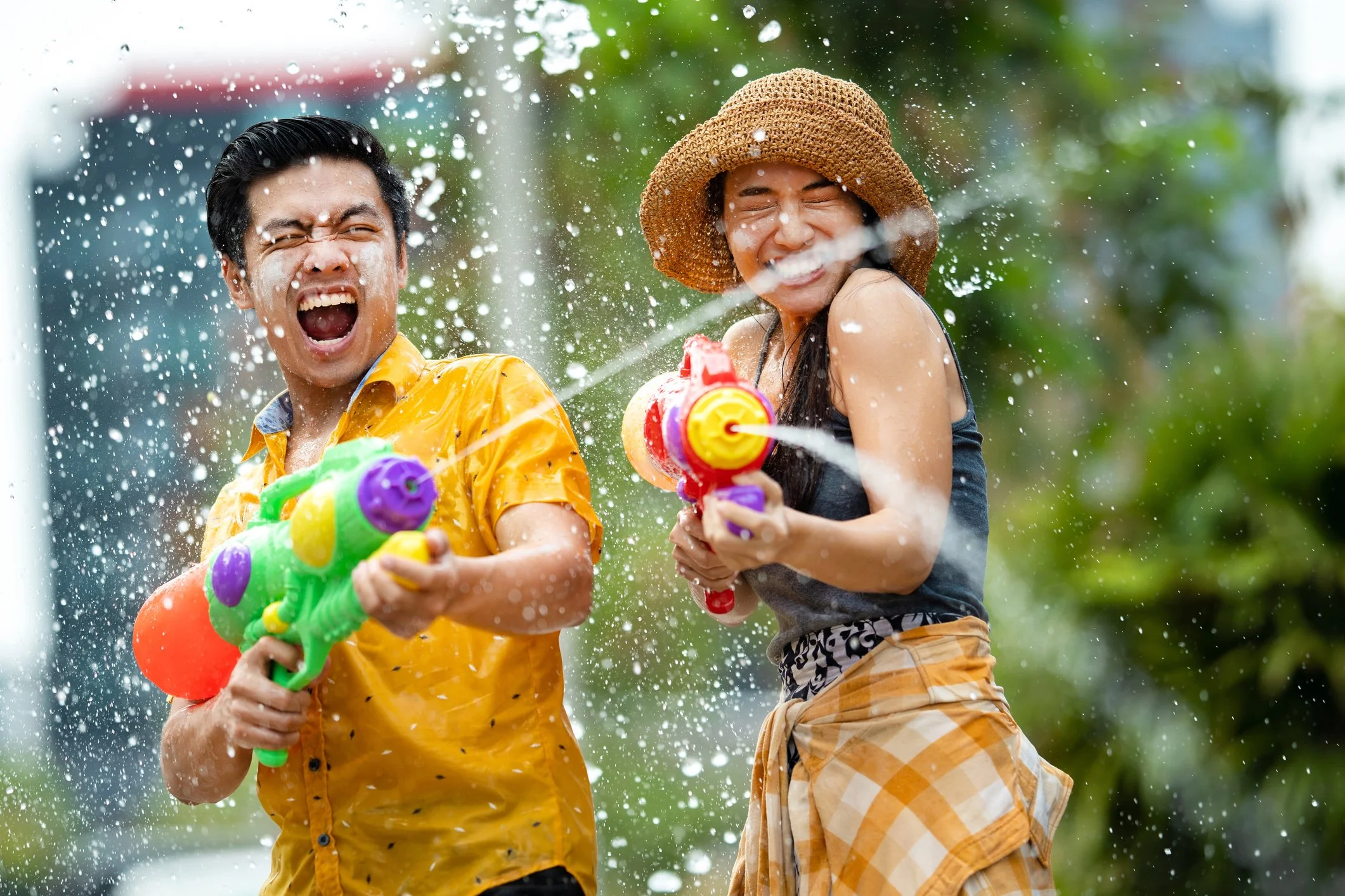 Two people smiling and laughing while playing with water guns during a water fight outside on a sunny day.