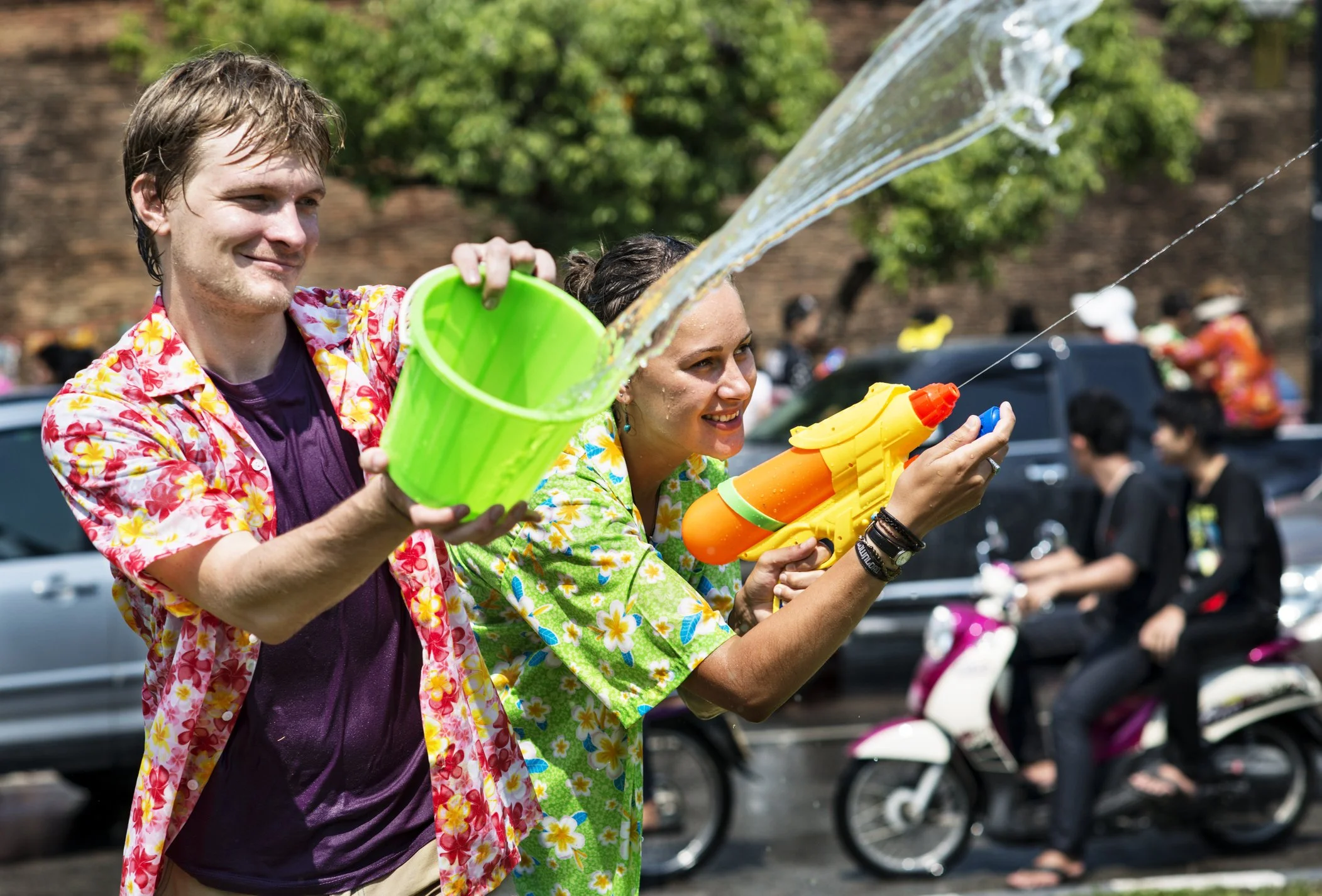 Two people in colorful Hawaiian shirts are playing with water guns on a street, with cars and a motorcycle in the background.