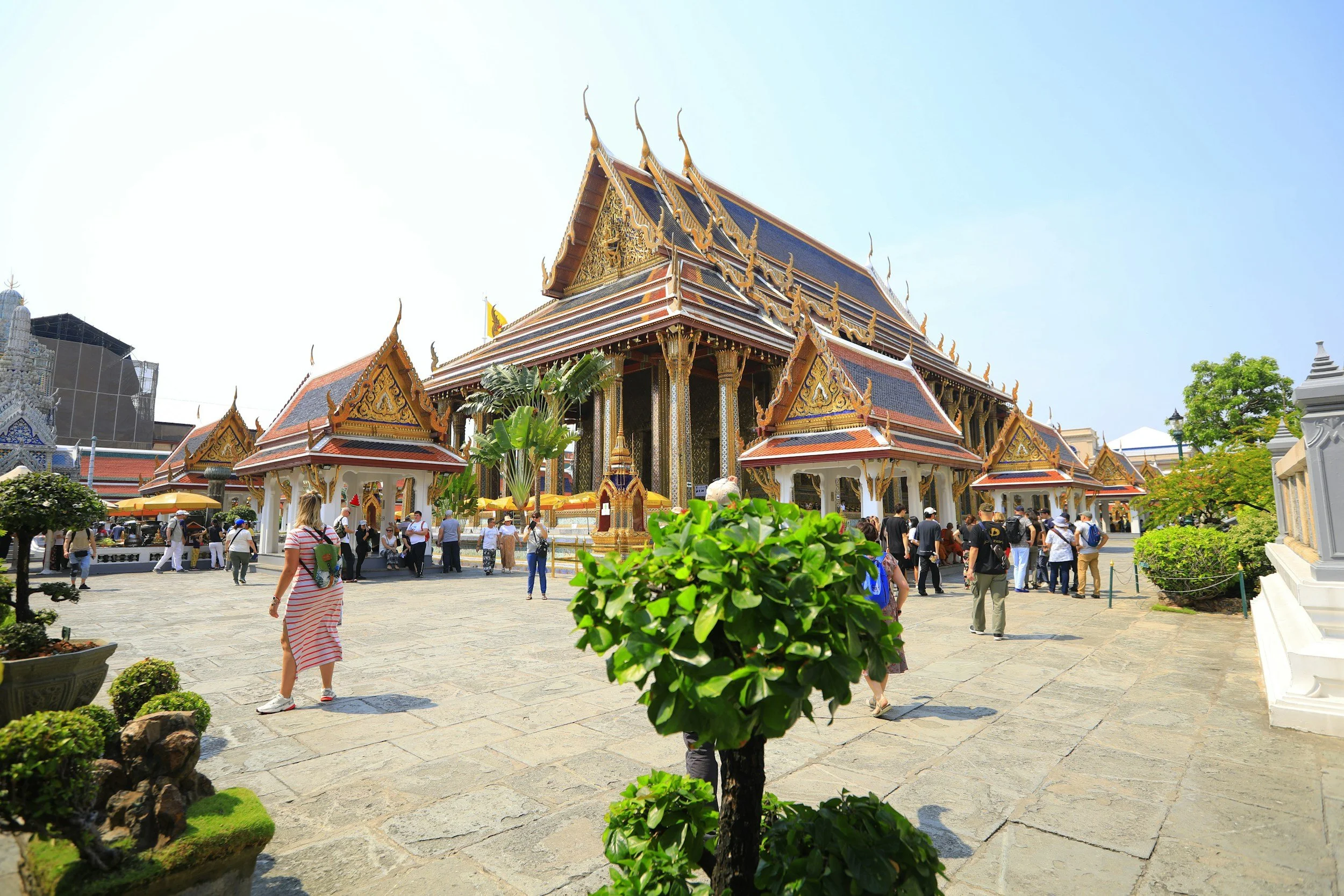 A traditional Thai temple with ornate gold and red roof details, surrounded by visitors and lush plants on a sunny day.
