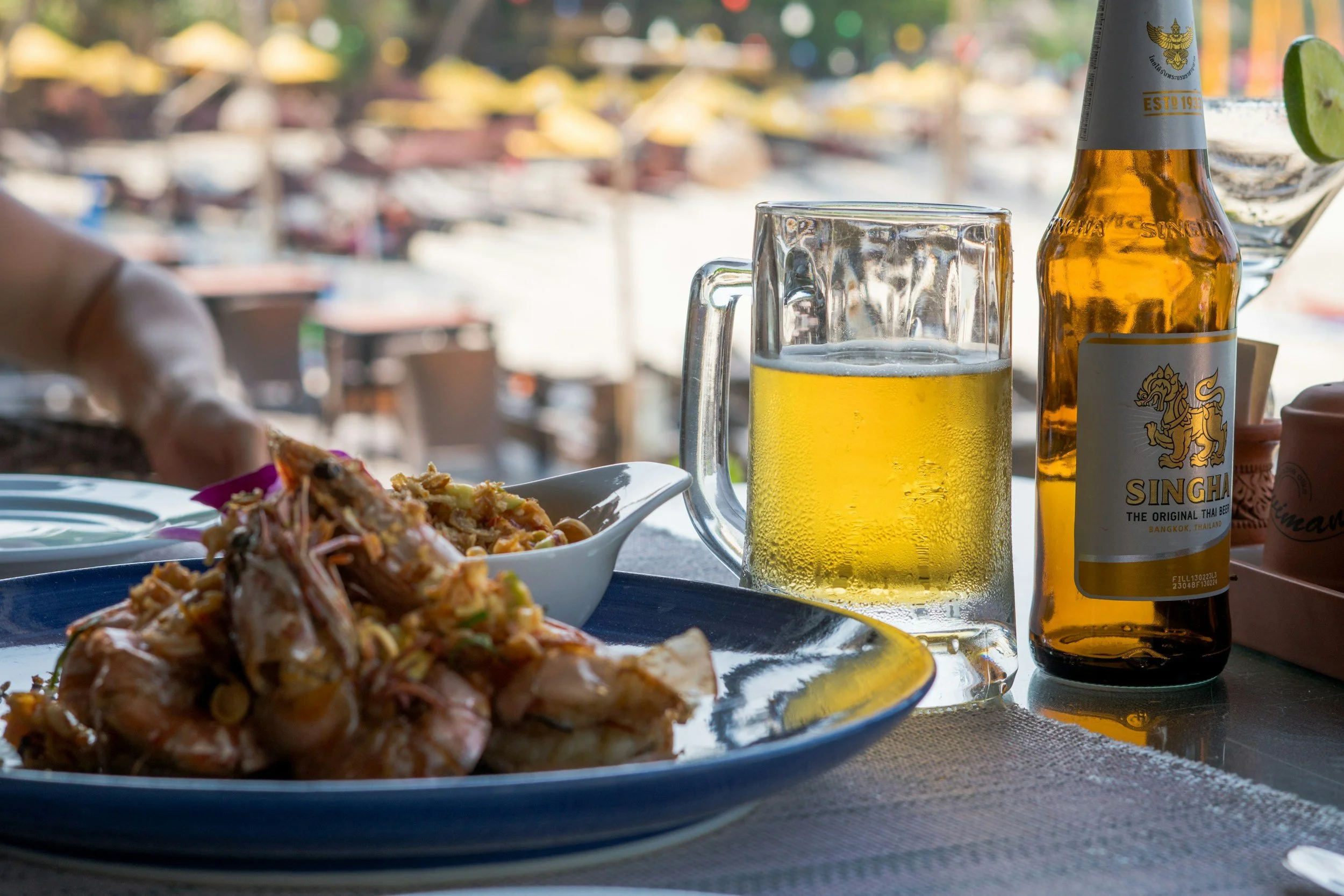 A dining table with a plate of food, a glass mug of beer, and a bottle of Singha beer on it, with outdoor seating in the background.