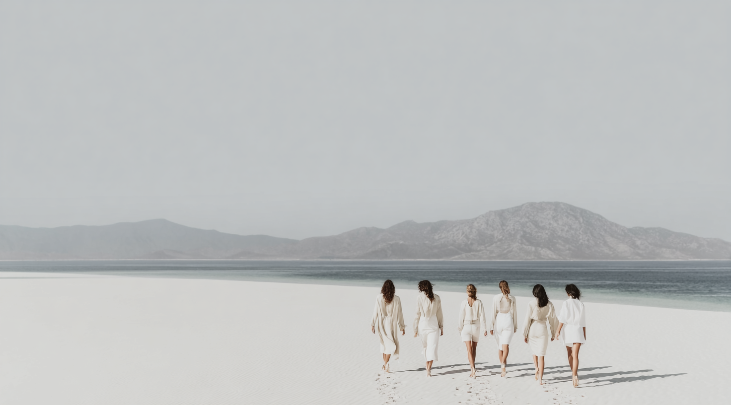 Seven women walking barefoot on white sand beach towards water with mountains in background.