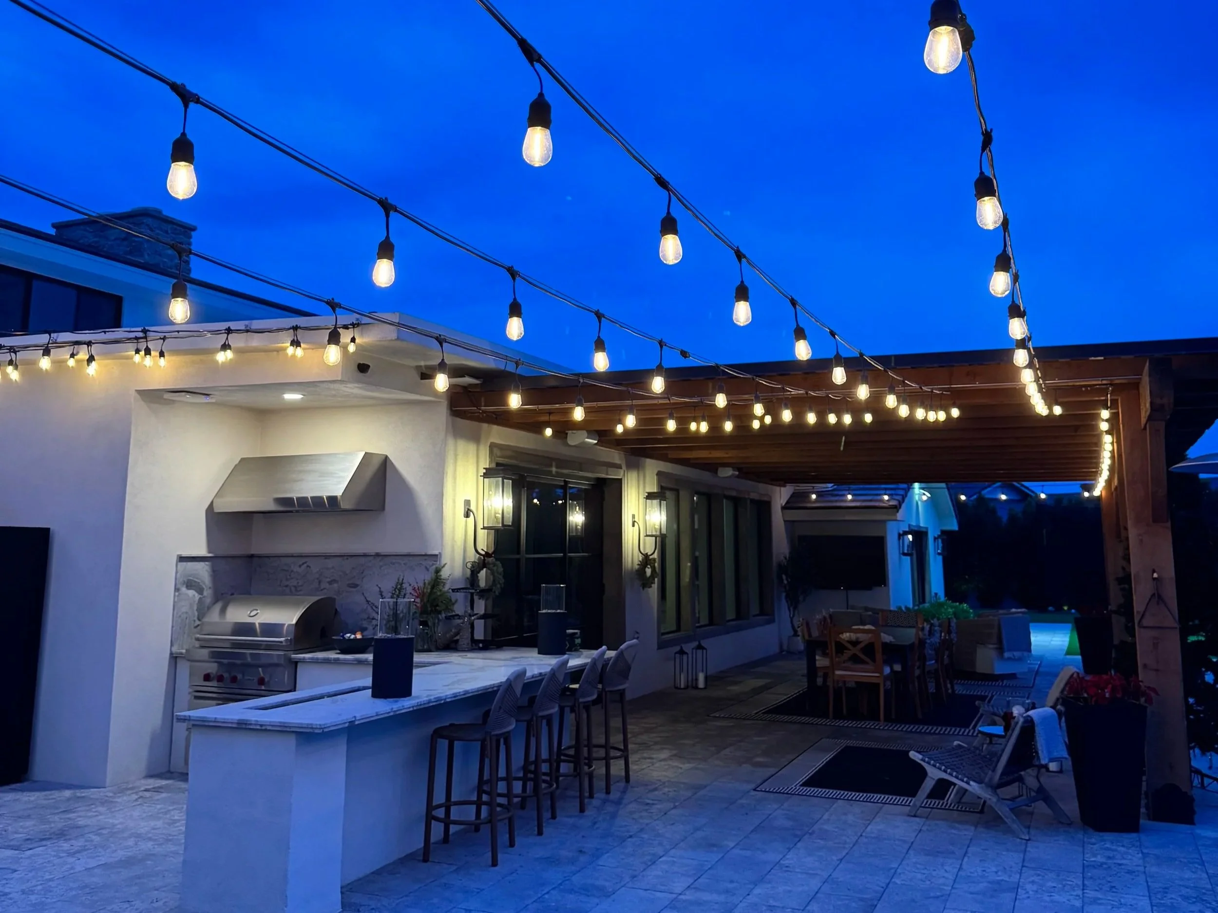 backyard patio with outdoor kitchen area, pergola and bistro lights at dusk with bright blue sky