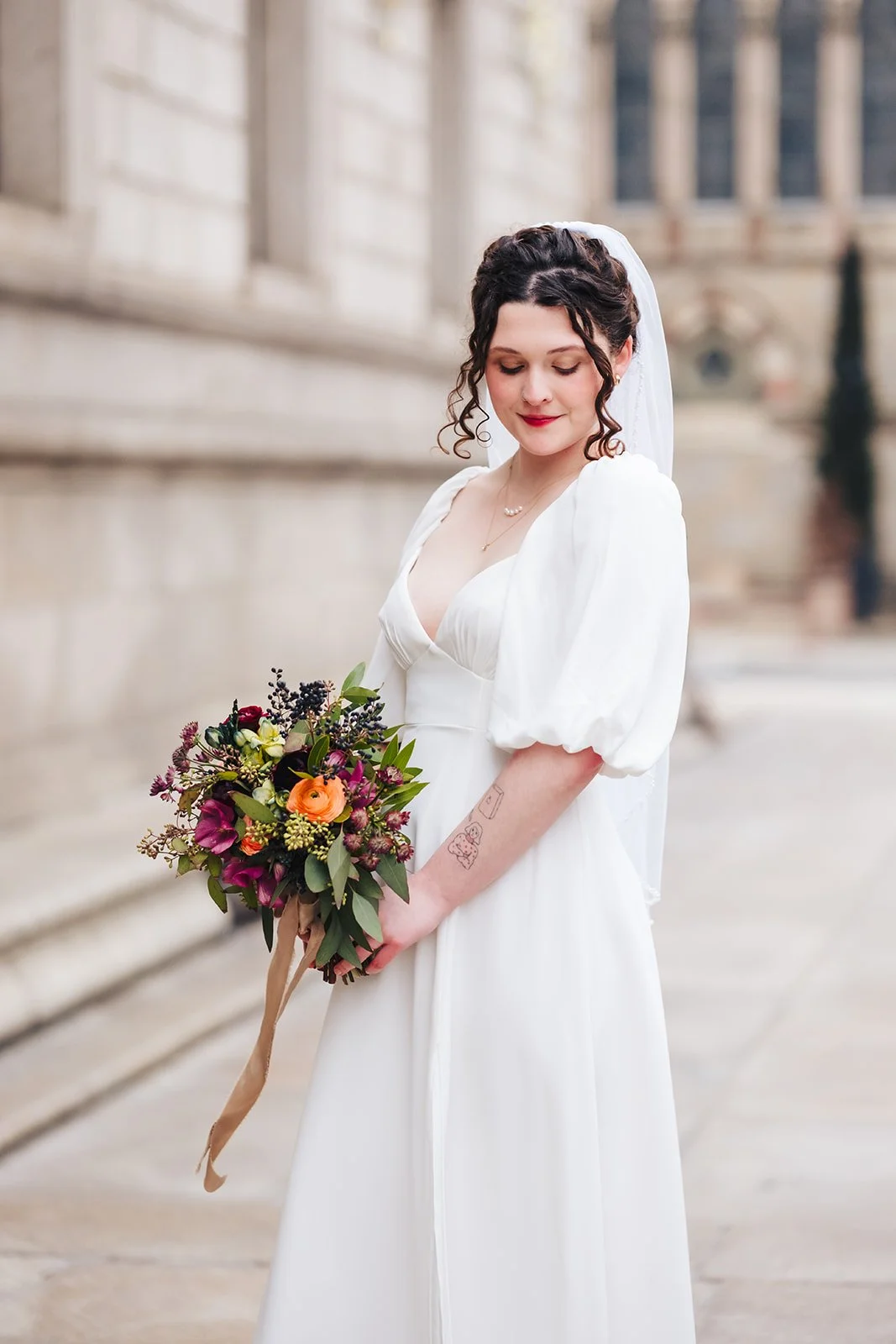 A bride in a white wedding dress holding a colorful bouquet of flowers, standing outdoors in front of an old stone building.